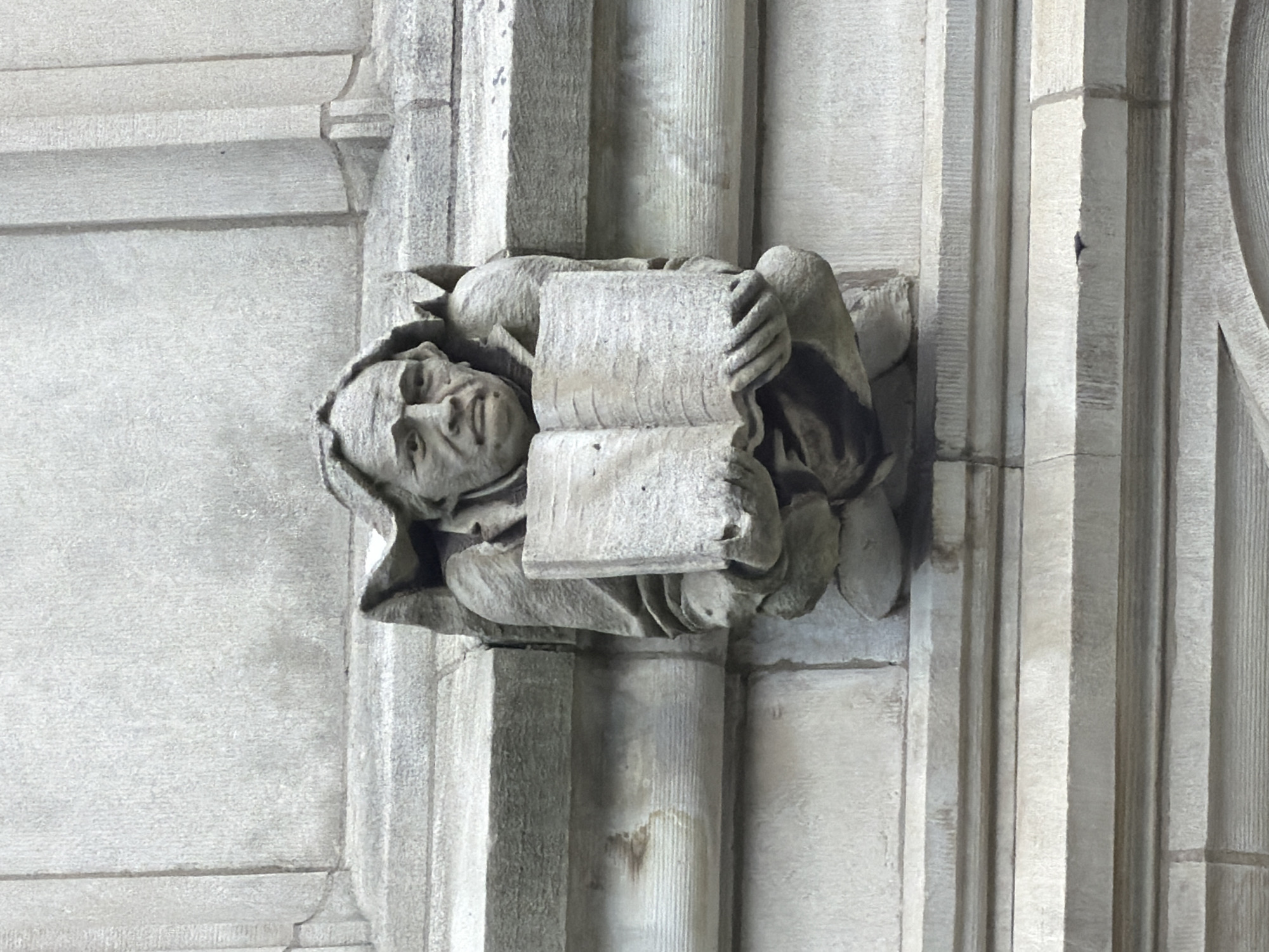A carved stone gargoyle, depicting a man with a furrowed brow and a stern expression, is affixed to the side of a building. The gargoyle is holding an open book with both hands. The figure is made of rough-hewn stone, and the details of its face, clothing, and the book are visible, though somewhat weathered. The background consists of the building's stone facade, characterized by horizontal bands and vertical lines, suggesting a classical architectural style. This gargoyle is located on the exterior of Firestone Library at Princeton University in Princeton, New Jersey, United States of America.