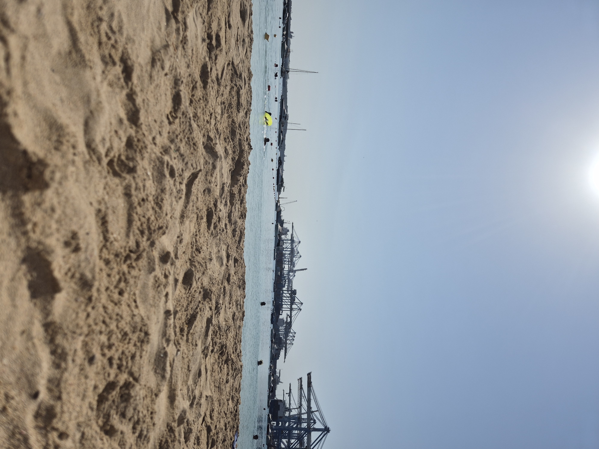The image depicts a sunny day at a beach in Birżebbuġa, Malta, with the foreground showing detailed textures of sand. In the middle ground, the clear blue sea is populated by several people swimming and one person on a yellow float, along with various boats and buoys. The background reveals a distant industrial port with numerous large cranes, characteristic of the Marsaxlokk Freeport, a significant maritime facility in Malta. The sky is bright blue with a faint sun visible.