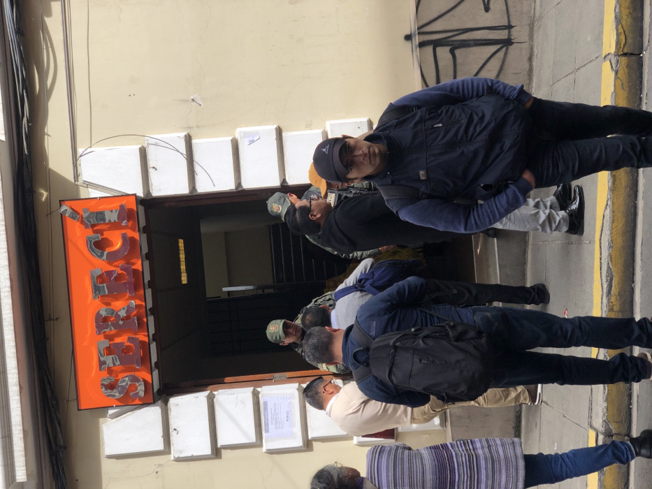 A group of people stands near a building with an orange sign that reads "SERVIC". Some wear hats and jackets, and one has a backpack. Several papers are affixed to the building's facade beneath the sign. The setting appears to be an urban area with a sidewalk adjacent to the building. The photo's perspective is slightly tilted.
