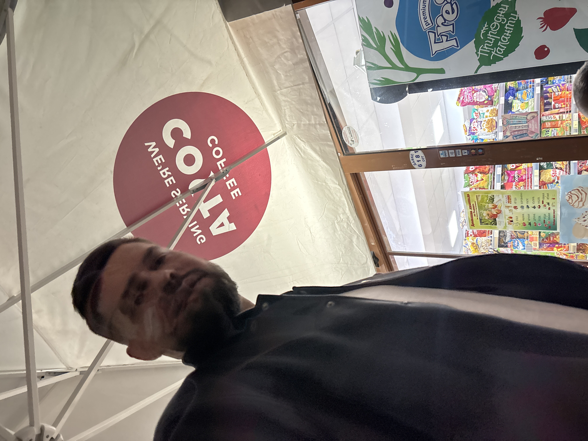 The image shows a man with a beard and dark hair standing under a Costa Coffee branded tent in front of a shop. The shop has a refrigerator section visible with a variety of snacks and drinks. The location is suggested to be in Plovdiv, Bulgaria, based on the presence of Bulgarian text on the advertisements and product packaging in the shop. The man is looking at the camera.