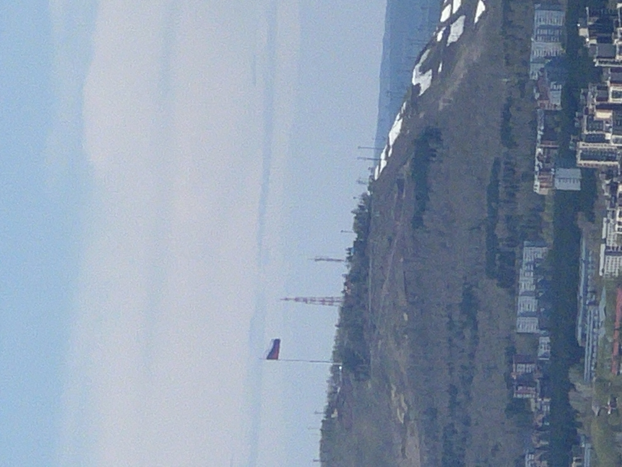 The image depicts an elevated view of a section of Krasnoyarsk, Russia, likely taken from a high vantage point. In the midground, a hill covered with vegetation slopes towards the foreground. A flag on a pole is visible on the hill, near the lower left of the frame. Behind the hill, there is a radio tower. Along the right side of the image, a dense collection of buildings, including residential apartments, form the cityscape. The sky is partially obscured by a haze.