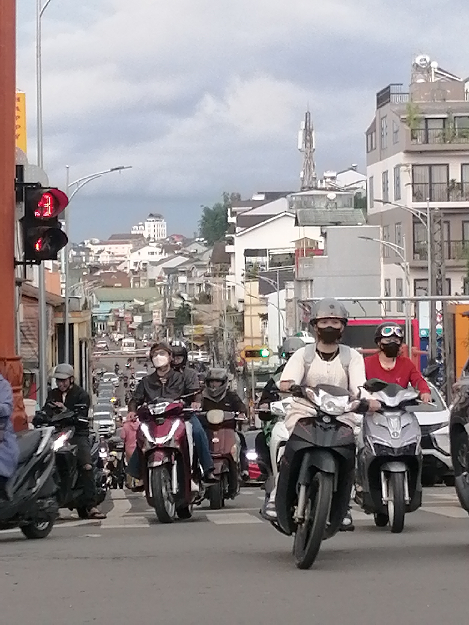 This is a street scene in Cam Ly - Đà Lạt, Vietnam, featuring a busy intersection with numerous motorcycles and pedestrians. A traffic light is visible on the left, displaying a red signal. Many individuals are riding motorcycles, most wearing helmets and face masks. The architecture in the background suggests a densely populated urban area with multi-story buildings and some hills. The overall atmosphere is one of moderate traffic and daily urban life.