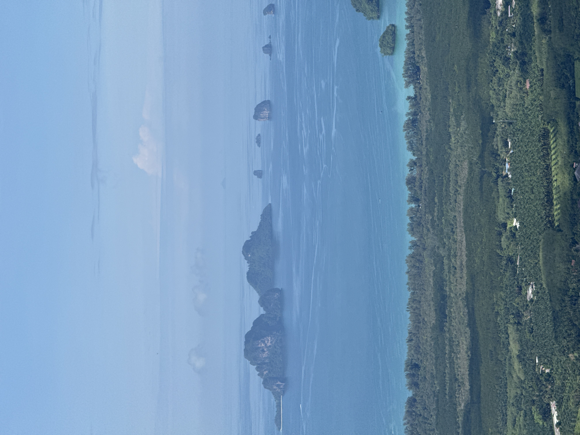 The image shows a panoramic view of the coastline near Ban Din Daeng Nui, Thailand. In the foreground, a dense forest covers the land, with some buildings scattered within. The forest transitions to a strip of trees along the coast, which meets a vast expanse of blue ocean. The ocean is dotted with several karst islands, some of which are covered in vegetation. The sky is a pale blue with a few scattered clouds. The overall scene is tranquil and natural, showcasing the tropical landscape of the region.