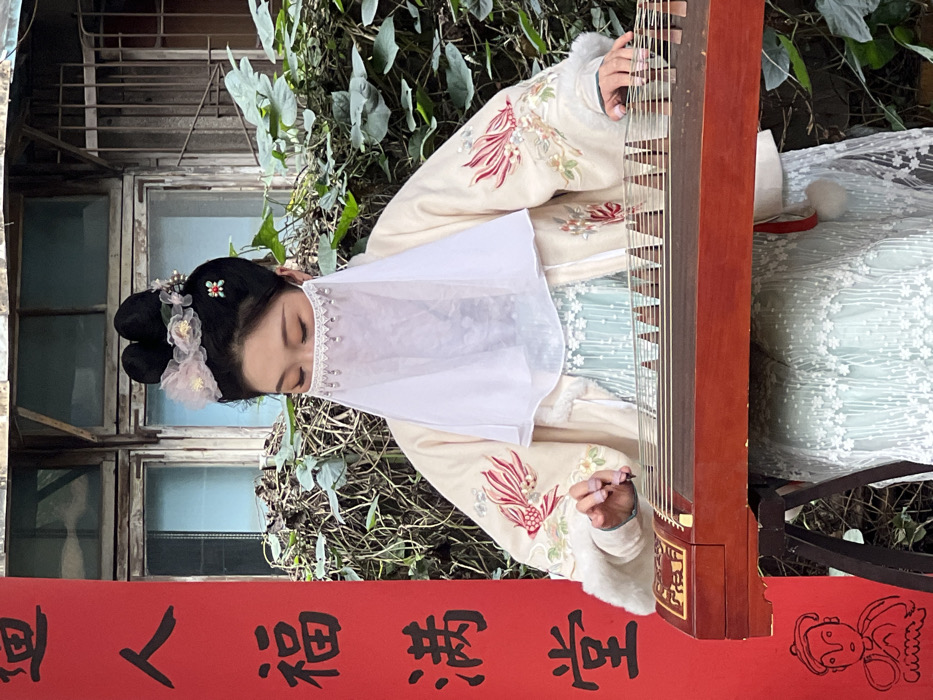 A woman in traditional garb plays a zither outdoors. She wears a cream-colored embroidered jacket and a white beaded veil. Her hair is styled with flowers. The instrument, a dark wood, is perpendicular to a red banner with black Chinese characters. Behind her are green foliage and a building with a weathered window. The shot is close-up, capturing the performer and her surroundings in detail.