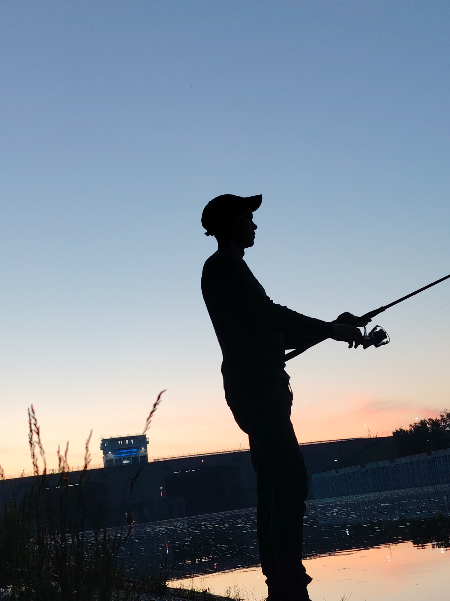 A silhouette of a man fishing on a body of water at dusk. The man is wearing a cap and holding a fishing rod. In the background, a dam with a structure on top is visible. The structure has writing on it, identifying it as located in Chaikovsky, Russia. The sky transitions from blue to orange. Grass and water are in the foreground.