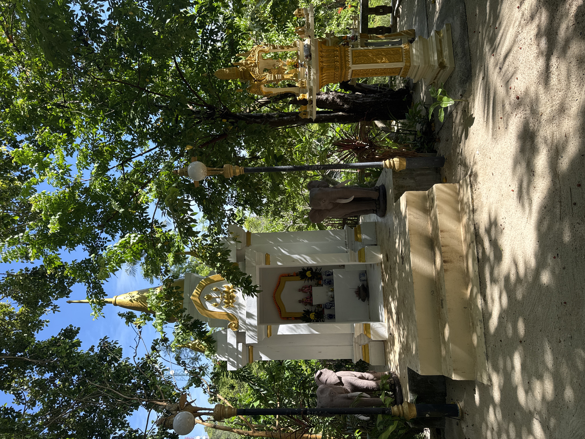 In the village of Koh Phangan, Thailand, a small, ornate shrine stands amidst lush greenery. The shrine is white with gold trim and features a tiered roof topped with a golden spire. In front of the shrine, two steps lead up to a platform. To the left of the shrine, a stone elephant statue stands guard. To the right, a taller, golden structure with multiple tiers and intricate carvings serves as another altar. The area is paved with a sandy-colored material, and dappled sunlight filters through the dense foliage overhead. The scene appears to be an outdoor religious site, possibly a Buddhist temple or a spirit house, reflecting the spiritual landscape of Thailand.