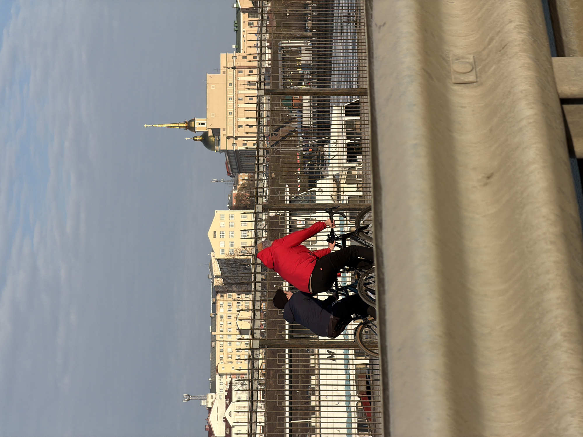 The image, taken from a high vantage point, shows two individuals on bicycles on a bridge, with the cityscape of Izhevsk, Russia in the background. A roadside barrier is in the foreground, obscuring the view to some extent. One person wears a red jacket, and the other a dark blue jacket. Buildings, including one with a golden dome and spire, are visible in the background. The sky is overcast. The scene depicts an urban environment with people engaged in recreational activity.