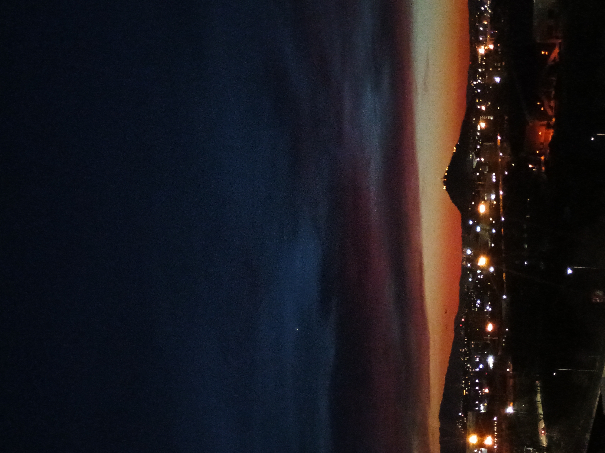 The image is a nighttime view of Plovdiv, Bulgaria, taken from an elevated position. To the left, the sky is dark blue. Transitioning towards the right, the sky shifts in color to a band of reddish-orange hues, likely from the setting sun or city lights. A mountain is visible in silhouette on the right side of the sky, forming the backdrop for the city. A line of lights stretches across the lower right portion of the image, indicating a cityscape. The lights include streetlights and building lights, depicting an urban area with varying levels of illumination.