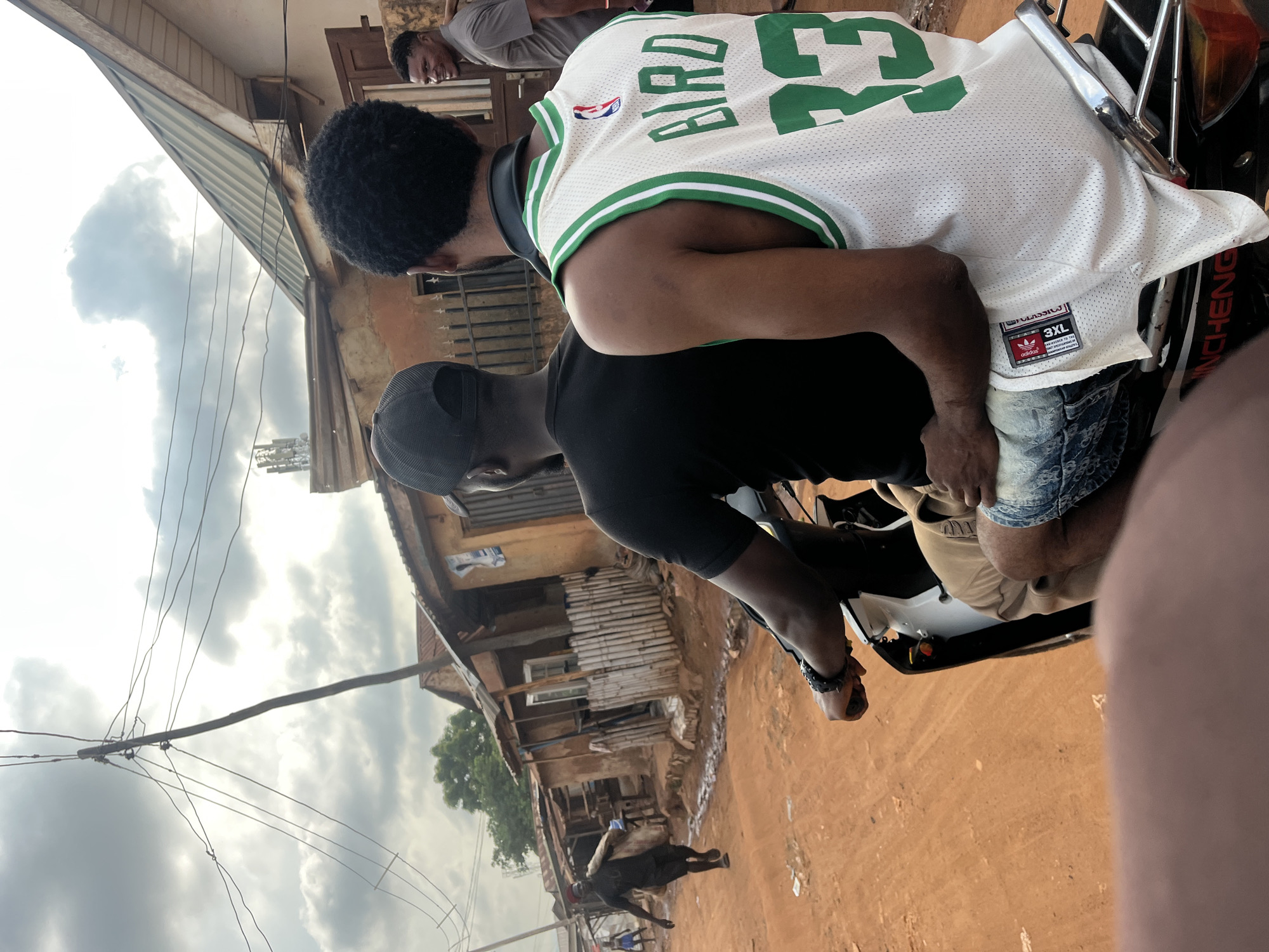The image captures a vibrant street scene in Abuja, Nigeria, focusing on two men riding a motorcycle. The passenger, viewed from behind, wears a white basketball jersey with "BIRD 33" prominently displayed in green on the back. A visible tag on the jersey indicates it's a "CLASSICS" item, size "3XL," featuring the Adidas and NBA logos. He has dark, curly hair and is wearing blue denim shorts. The driver, mostly obscured, is dressed in a black t-shirt and a black baseball cap, operating a motorcycle with "QINCHENG" visible on its side. Another man in a grey t-shirt is seen standing in the background behind the passenger.
The setting is an unpaved, reddish-brown dirt road, indicative of laterite soil common in the region. Along the street, several simple, rustic buildings are visible, constructed from materials like wood, corrugated metal, and possibly concrete or mud bricks, suggesting a local residential or commercial area. One building has a corrugated metal awning. Overhead, a wooden utility pole supports multiple sagging power lines against a bright, partly cloudy sky. The time of day appears to be daytime, likely late morning or afternoon, under an overcast or scattered cloud cover. In the distance, other individuals are visible on the street, contributing to the everyday atmosphere of the scene.