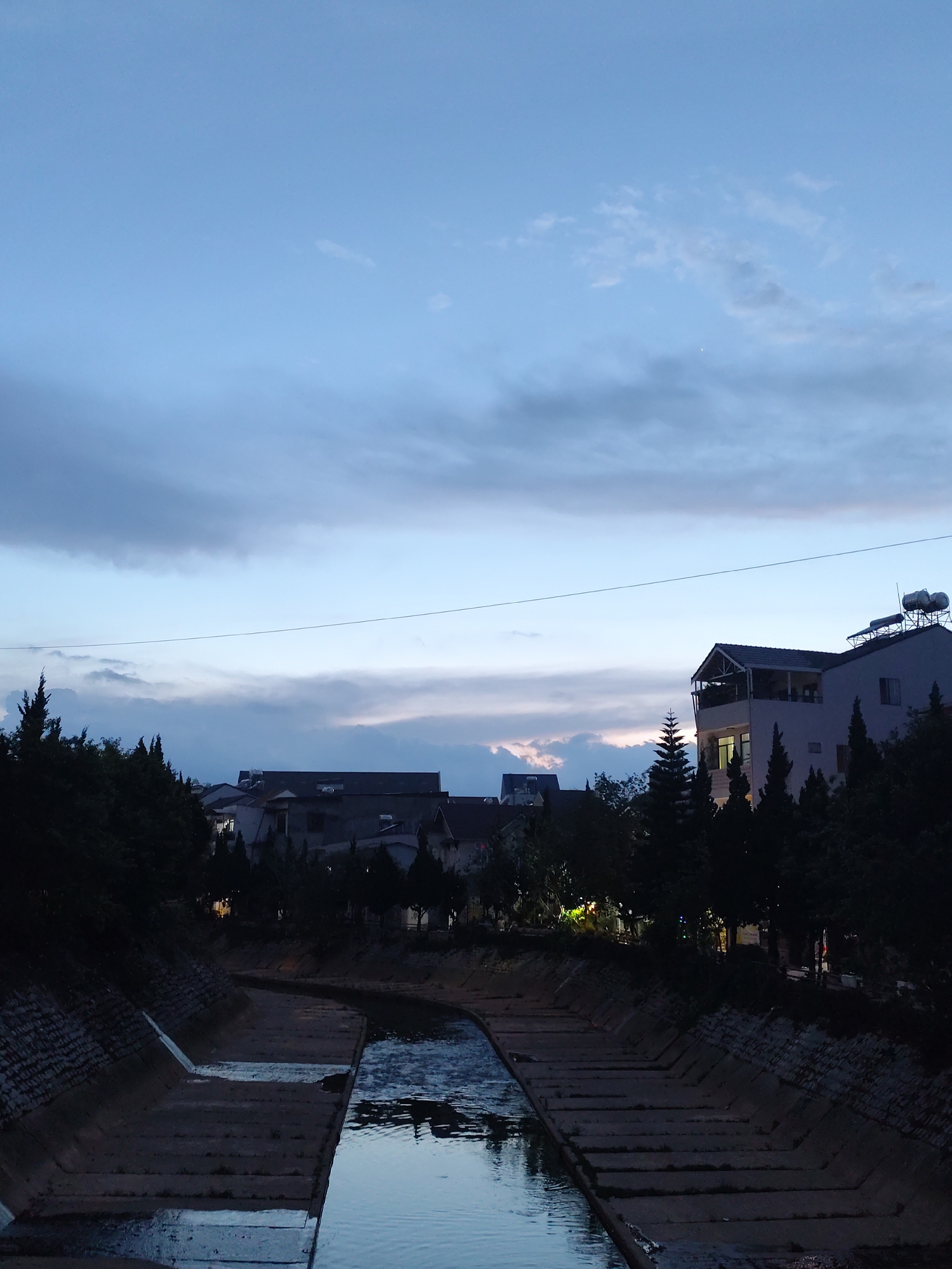 The image captures a tranquil twilight scene over a concrete-lined canal in what appears to be Da Lat, Vietnam. The sky, transitioning from day to night, dominates the upper portion, displaying a gradient of deep blue at the zenith fading into lighter blues, soft pinks, and oranges near the horizon where the last vestiges of sunset glow. Scattered grey and white clouds drift across the sky, and a single bright point of light, possibly a star or planet, is visible towards the top. A thin power line horizontally dissects the upper-middle frame.
Below the sky, the landscape features a mix of urban development and natural elements. Dark, silhouetted evergreen trees, possibly pines or cypress, line both banks of the canal and extend behind the buildings, their sharp forms contrasting with the fading light. Various multi-story buildings, likely residential or commercial structures, are visible amongst the trees. Some buildings feature balconies and visible windows, with a few showing internal lights, indicating the progression into evening. Several rooftops are equipped with solar water heaters.
The concrete-lined canal flows through the center of the lower frame, its calm water reflecting the soft sky colors. The canal banks are constructed from concrete blocks and stone, showing some small patches of vegetation. No people are visible, and no specific activity is taking place, contributing to a serene and quiet atmosphere. There is no visible text in the image.