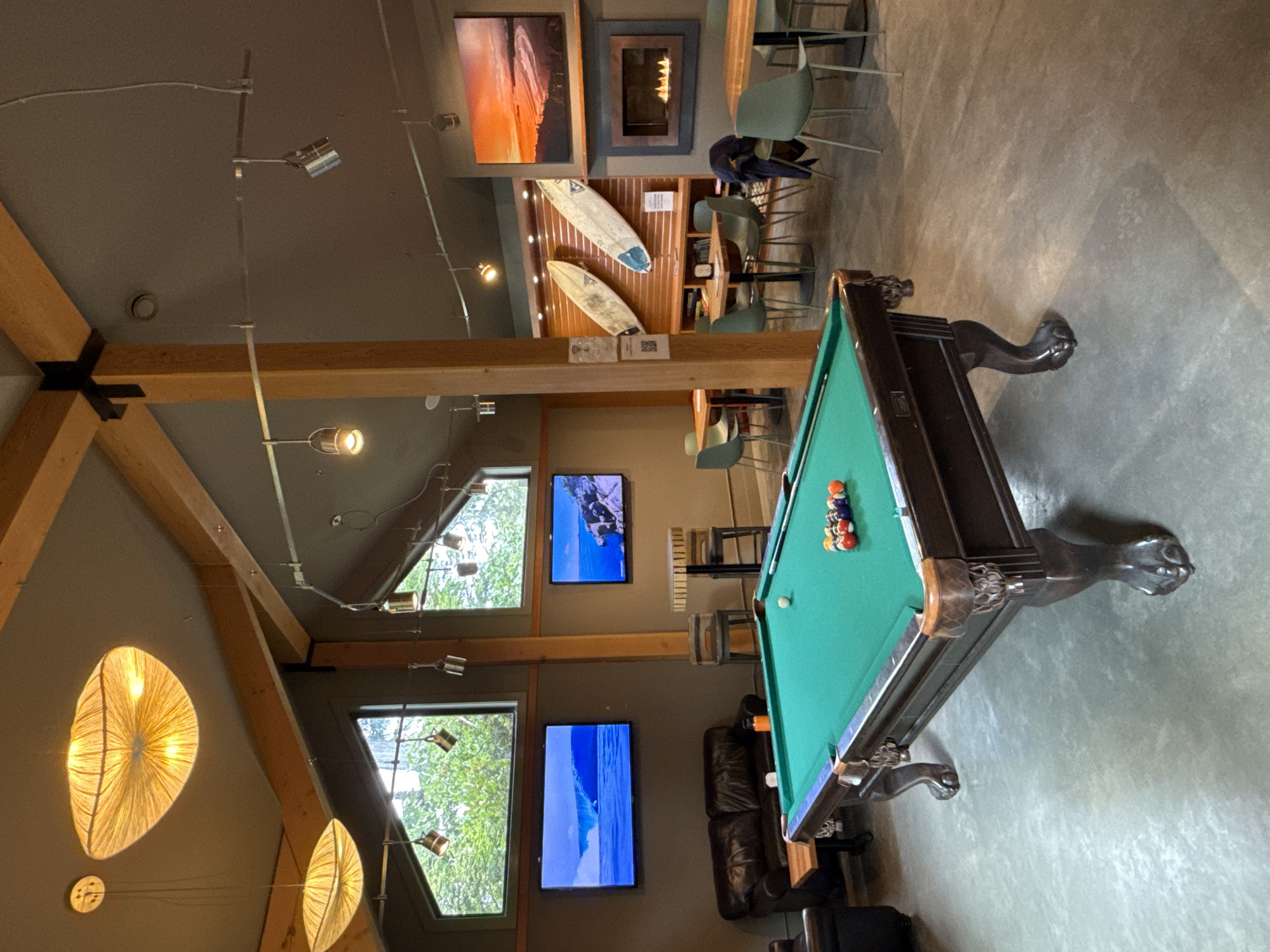 The image depicts an indoor recreational area, likely a common space in accommodation in Tofino, Canada, as indicated by the surfboards displayed on the wall above a fireplace. A pool table dominates the right side of the frame, balls racked at one end. Adjacent to the pool table is a leather sofa. Two televisions hang on the wall above the sofa. Large windows are present, displaying greenery and natural light. Distinctive lighting fixtures hang from the ceiling. Furniture includes various chairs and tables placed near the fireplace.