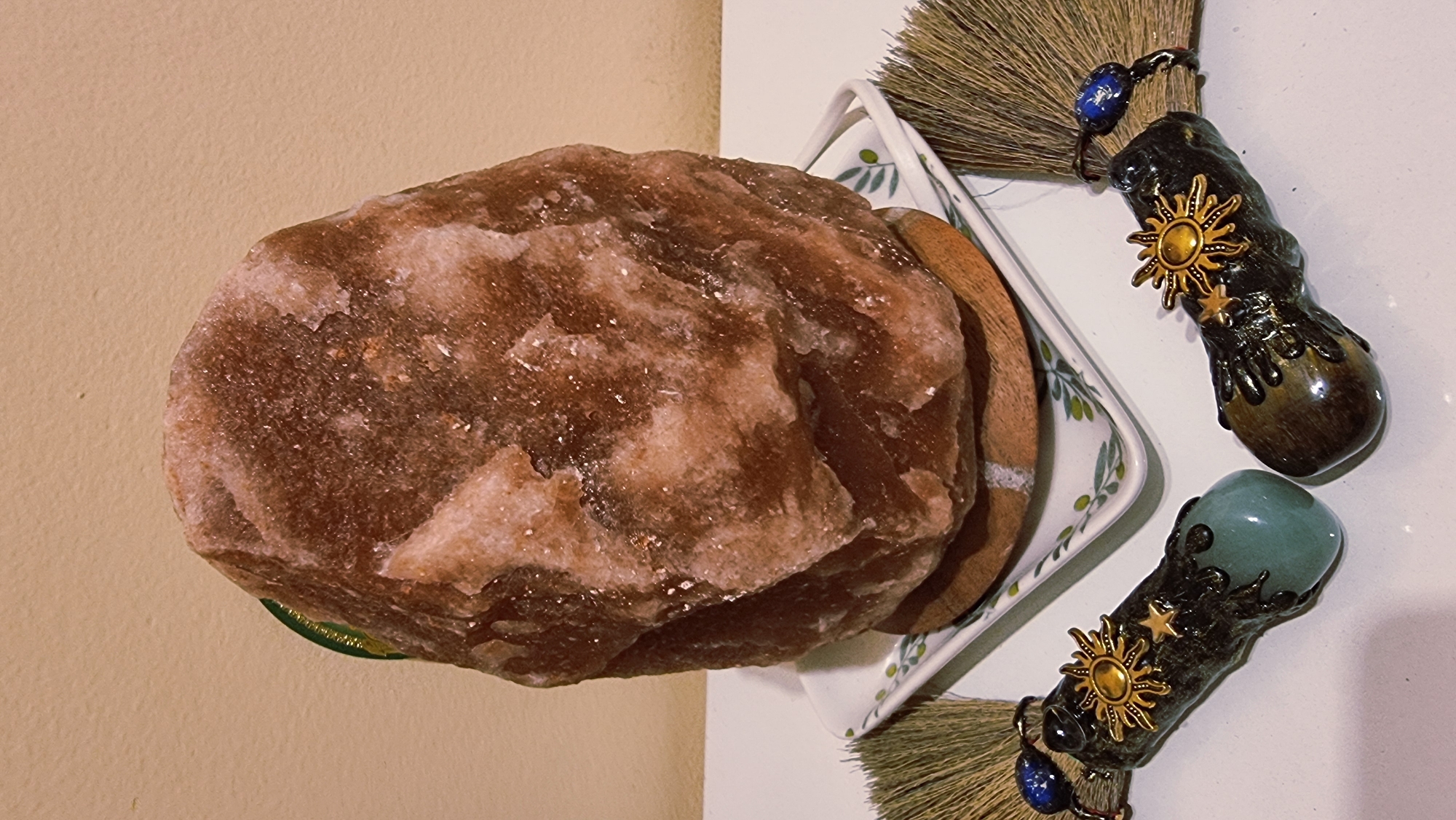 This is a close-up, eye-level, indoor photograph of a large, irregular, reddish-brown Himalayan salt lamp resting on a small white square dish with a green olive branch pattern. The dish is placed on a wooden base. Flanking the salt lamp are two small, decorative brooms with dark, textured handles. The broom on the left has a smooth, light green gemstone at its base and is adorned with a golden sunburst and stars. The broom on the right has a darker, mottled gemstone at its base and is similarly decorated with a golden sunburst and stars. Both brooms have natural, straw-colored bristles, and a small blue bead is visible on the bands holding the bristles. The background is a plain, light beige wall. The lighting is warm and appears to come from the front, casting soft shadows. The scene is static and suggests a display of decorative or ritualistic objects, possibly in a home or shop. There are no people visible, and no actions are taking place. The photograph does not provide any information to determine its location in Sant Joan Les Fonts, Spain.