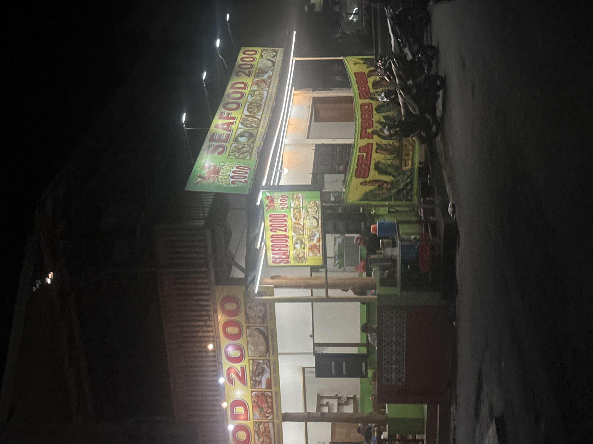 The image captures a nighttime view of a seafood restaurant called "SEAFOOD 2000" in the City of Binjai, Indonesia. The restaurant appears to be a casual establishment with a brightly lit facade. Several signs display the restaurant's name and images of seafood dishes. A banner hangs in front, also advertising "SEA FOOD 2000." A few people are visible inside and around the restaurant, and several motorcycles are parked on the side of the road in front of the establishment. The overall scene suggests a lively, local dining spot operating in the evening.