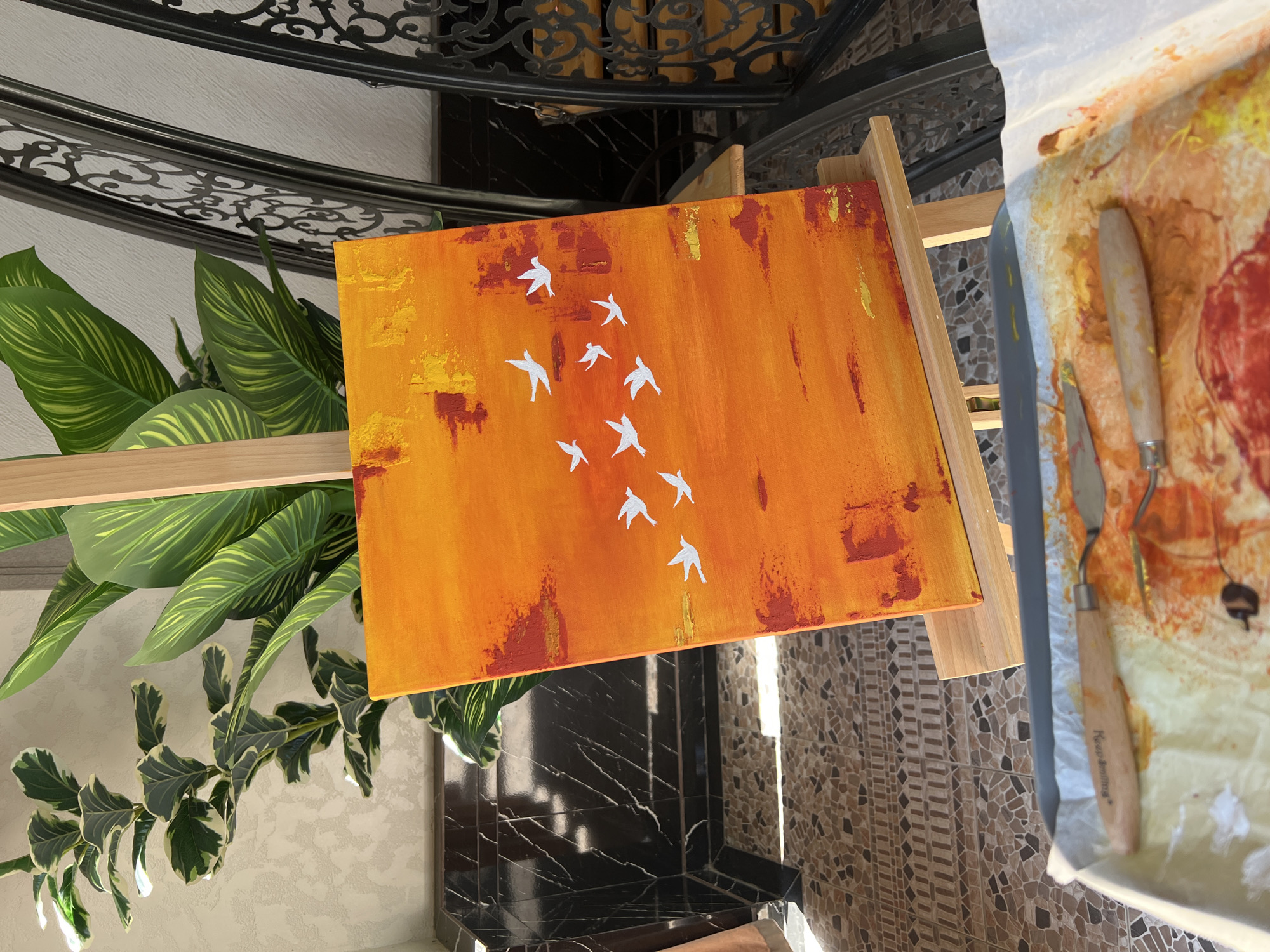 An abstract painting of white birds in flight against a vibrant orange and yellow background rests on a wooden easel. The painting is positioned indoors, near a window that casts a bright light, illuminating the textured brushstrokes. To the left of the easel, a large, leafy green plant stands, its variegated leaves catching the sunlight. In the foreground, a palette tray with paint-covered spatulas and paper suggests an ongoing art project. The background reveals a tiled floor and a black, ornate metal railing, possibly part of a staircase or balcony, hinting at an interior space with decorative architectural elements. The scene is set in Namangan City, Uzbekistan.