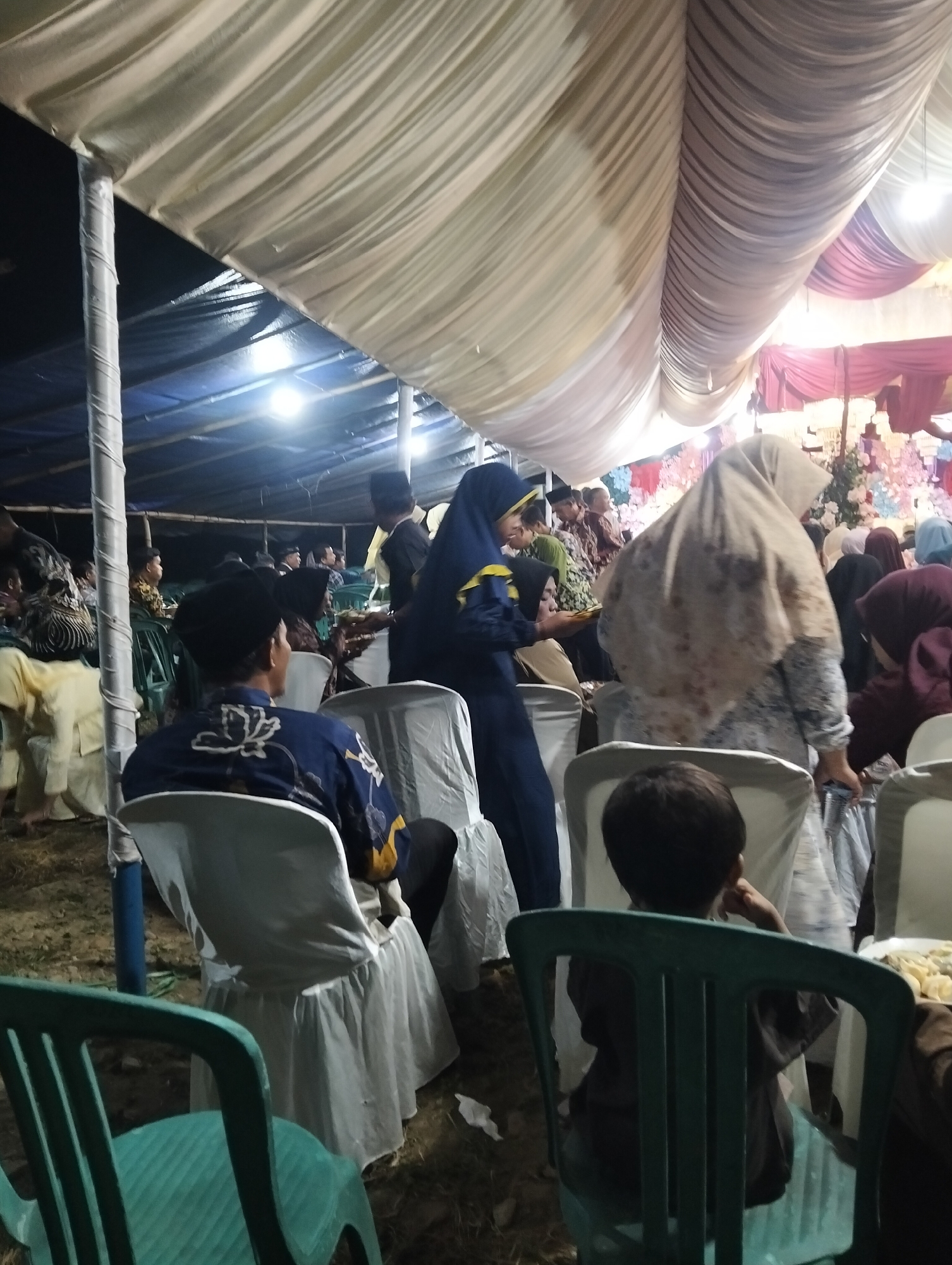 The image depicts an outdoor event, likely a wedding reception, taking place under a large tent in Bambalemo, Indonesia. The tent's ceiling is draped with white and maroon fabric, creating a festive atmosphere. Various individuals are seated on white chairs, some of which are covered with white fabric. In the foreground, a young boy is seated on a green plastic chair, facing away from the camera. To his left, a man in a blue shirt with white floral patterns is seated, also facing away. Further back, several women are visible, some wearing headscarves. The ground appears to be dirt. The lighting is dim, with some artificial lights visible within the tent.