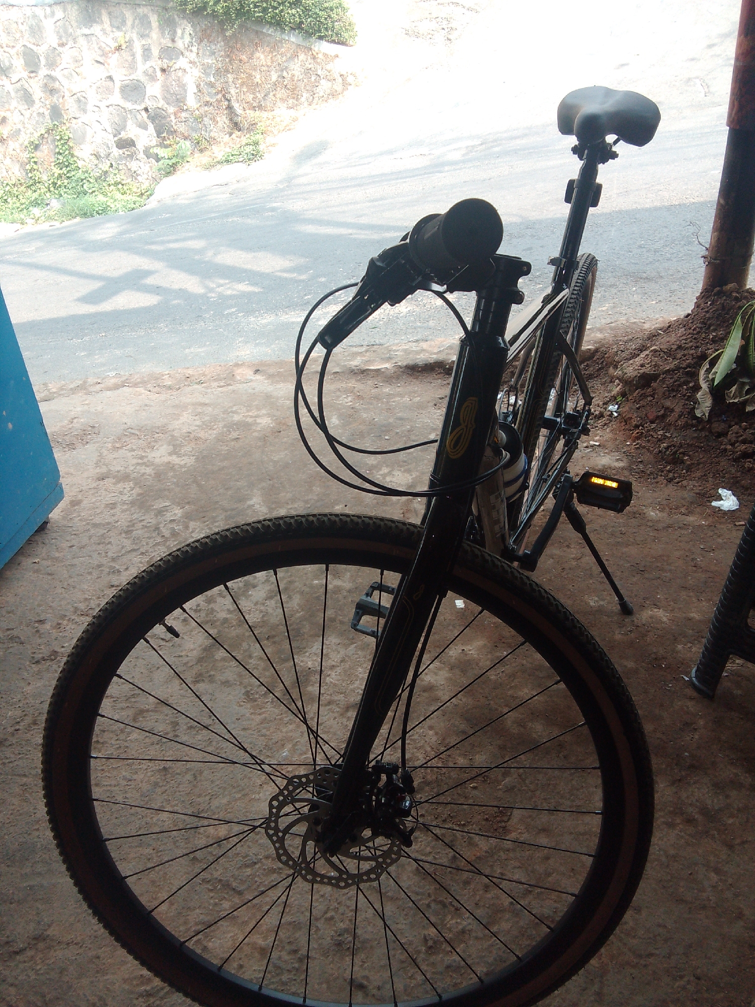 A black bicycle is parked outdoors on a dirt and concrete surface. The bicycle is oriented with its front wheel facing the camera and slightly to the left. It features a disc brake on the front wheel and a kickstand deployed on the right side, supporting it upright. The handlebars are visible, with black grips and cables. A black bicycle seat is mounted on a silver seat post. The frame of the bicycle is black with yellow detailing and a water bottle cage holding a white bottle.
The background shows a sloping asphalt road with shadows cast by sunlight. To the left of the bicycle, a portion of a blue structure is visible. Behind the road, there is a stone wall covered with vegetation. The ground is a mix of dirt and concrete, with some small white debris scattered around. To the right of the bicycle, there is a pile of brown soil and some green plants. The overall environment suggests an outdoor setting, possibly in a residential or less urbanized area, consistent with locations in Cisarua, Indonesia, which is known for its cooler climate and natural surroundings. The lighting indicates it is daytime, with bright sunlight. There are no people present in the image.