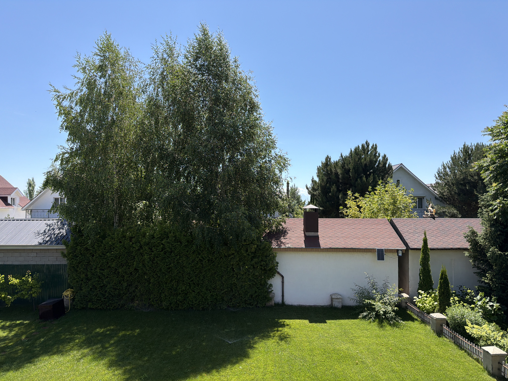 A bright, sunny day unfolds in this outdoor scene, showcasing a well-maintained backyard with a clear blue sky overhead. Dominating the left side of the frame is a large cluster of mature trees, their green foliage casting dappled shadows across the manicured lawn. Adjacent to the trees, a tall, dense hedge creates a privacy screen.
Behind the hedge, a low-slung building with a reddish-brown tiled roof stretches across the middle ground. A white wall forms the main facade of this structure, punctuated by a dark brown chimney. To the far right, more trees and lush greenery are visible, interspersed with glimpses of residential buildings in the background. A small, decorative fence runs along the right edge of the lawn, separating it from the denser vegetation.
In the foreground, the vibrant green grass appears to have been recently watered, with visible signs of a sprinkler system. A yellow garden hose is coiled near the hedge. The scene is peaceful and idyllic, suggesting a quiet day in a suburban or rural setting. The lighting and clear sky indicate it is daytime, likely late morning or early afternoon. No people or animals are visible, and there is no text present in the image.