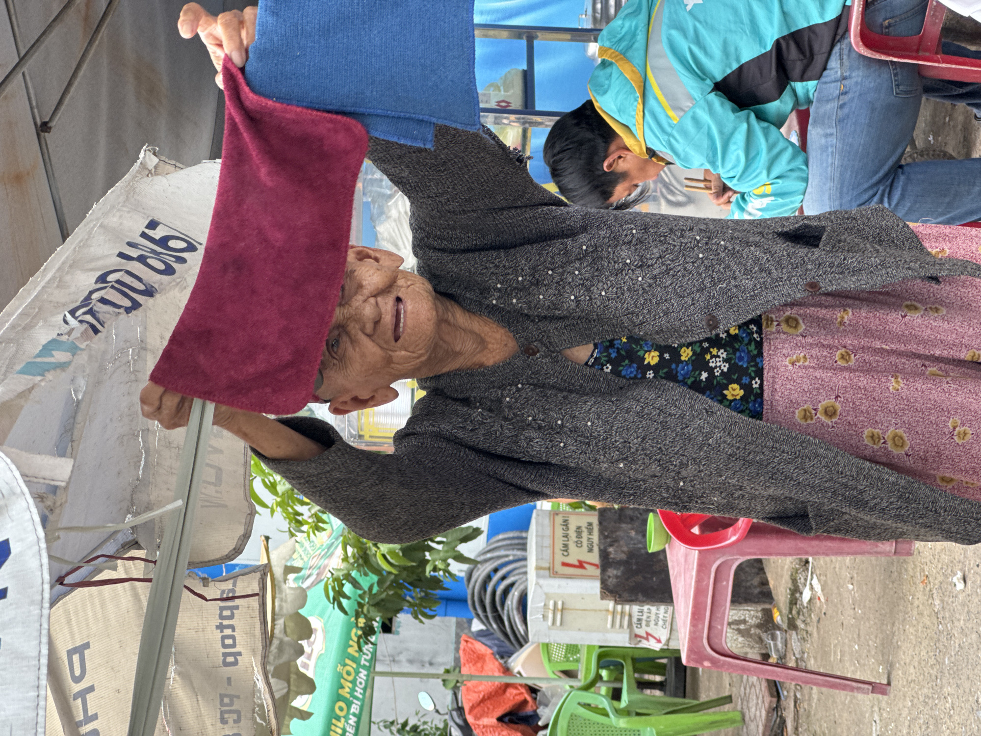 An elderly woman is standing outdoors, holding up a piece of maroon fabric and a blue fabric, with one hand on the maroon fabric and the other hand underneath the blue fabric. She is wearing a dark grey knitted cardigan over a floral-patterned shirt, and a pink skirt with a floral pattern. The woman has wrinkled skin and is smiling with her mouth open. Behind her, a young man in a bright turquoise jacket with yellow accents is sitting at a red table, holding chopsticks and eating. Various objects are visible in the background, including green plastic chairs, a red plastic table, coiled black hoses, white boxes with Vietnamese text, and a motorcycle. The scene appears to be in a market or street setting, with awnings and other temporary structures visible. The overall lighting suggests it is daytime.