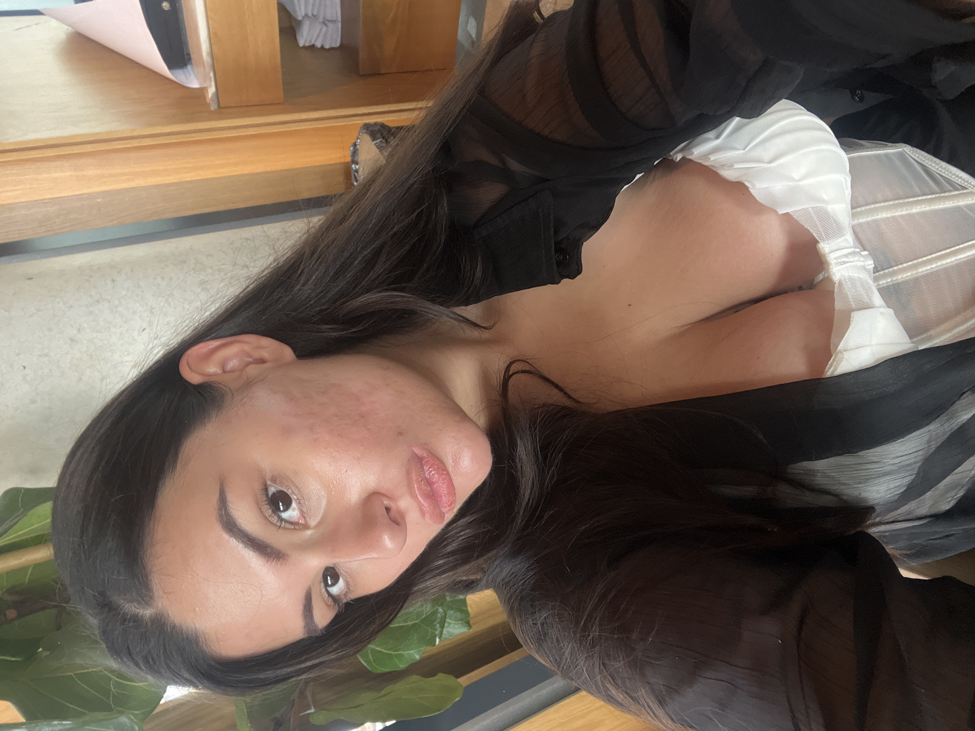 A woman is seen in a close-up shot. She has long dark hair and is wearing a white corset underneath a sheer black top. Her face is visible, displaying fair skin and noticeable skin texture on one side. The background is somewhat blurred, but reveals parts of wooden furniture or structure and green foliage. The location is not discernible from the image.