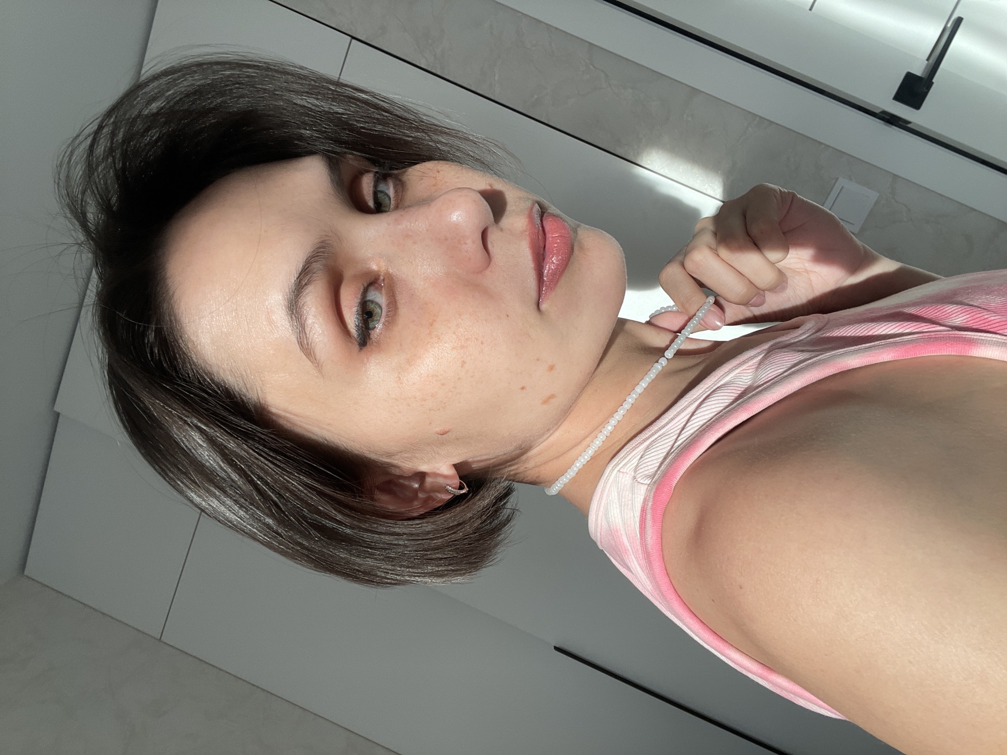 A head-and-shoulders angled portrait depicts a young woman with short, dark hair and fair skin. She holds a beaded necklace near her neck with her right hand. Her face shows freckles and a neutral expression. She is wearing a pink tank top. The background appears to be an interior space with a white or light-colored wall. The location is not discernible as Izhevsk, Russia, based on visual cues in the image.