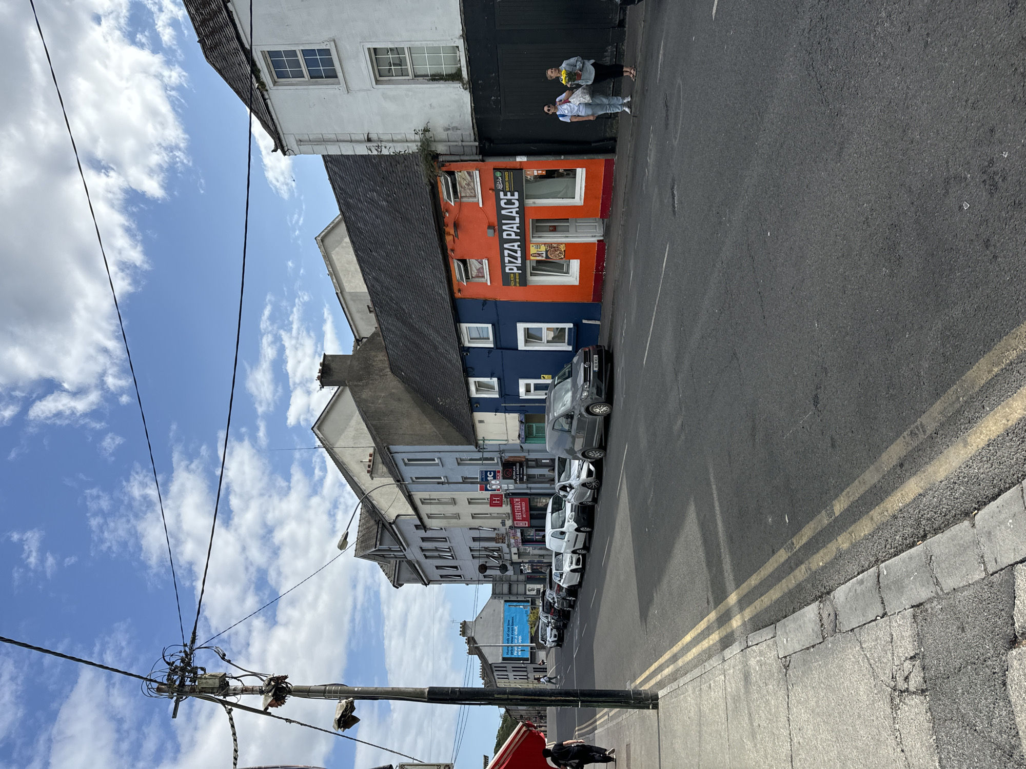 The image captures a street scene in Waterford, Ireland, likely on a sunny day with scattered clouds. The viewpoint is from the sidewalk, looking down a street lined with buildings. On the left side, a black utility pole with numerous wires and a lamp is prominent. A person in dark clothing walks away from the camera on this sidewalk.
The street itself is paved with asphalt and marked with double yellow lines. Several cars are parked along the right side of the street, including a silver van and a dark gray van parked at the end.
The buildings along the right side of the street are varied in color and architecture. The most prominent is a bright orange building with a sign that reads "PIZZA PALACE." Adjacent to it is a blue building with white-trimmed windows. Further down the street, there are buildings painted in various shades of gray, blue, and white, with signs for businesses like "HERTERICH AUCTIONEERS."
Two women are standing on the sidewalk in front of the "PIZZA PALACE." One is wearing a white top and blue jeans, holding a bouquet of yellow flowers. The other is wearing a dark top and pants. The overall atmosphere suggests a typical urban street with commercial activity.