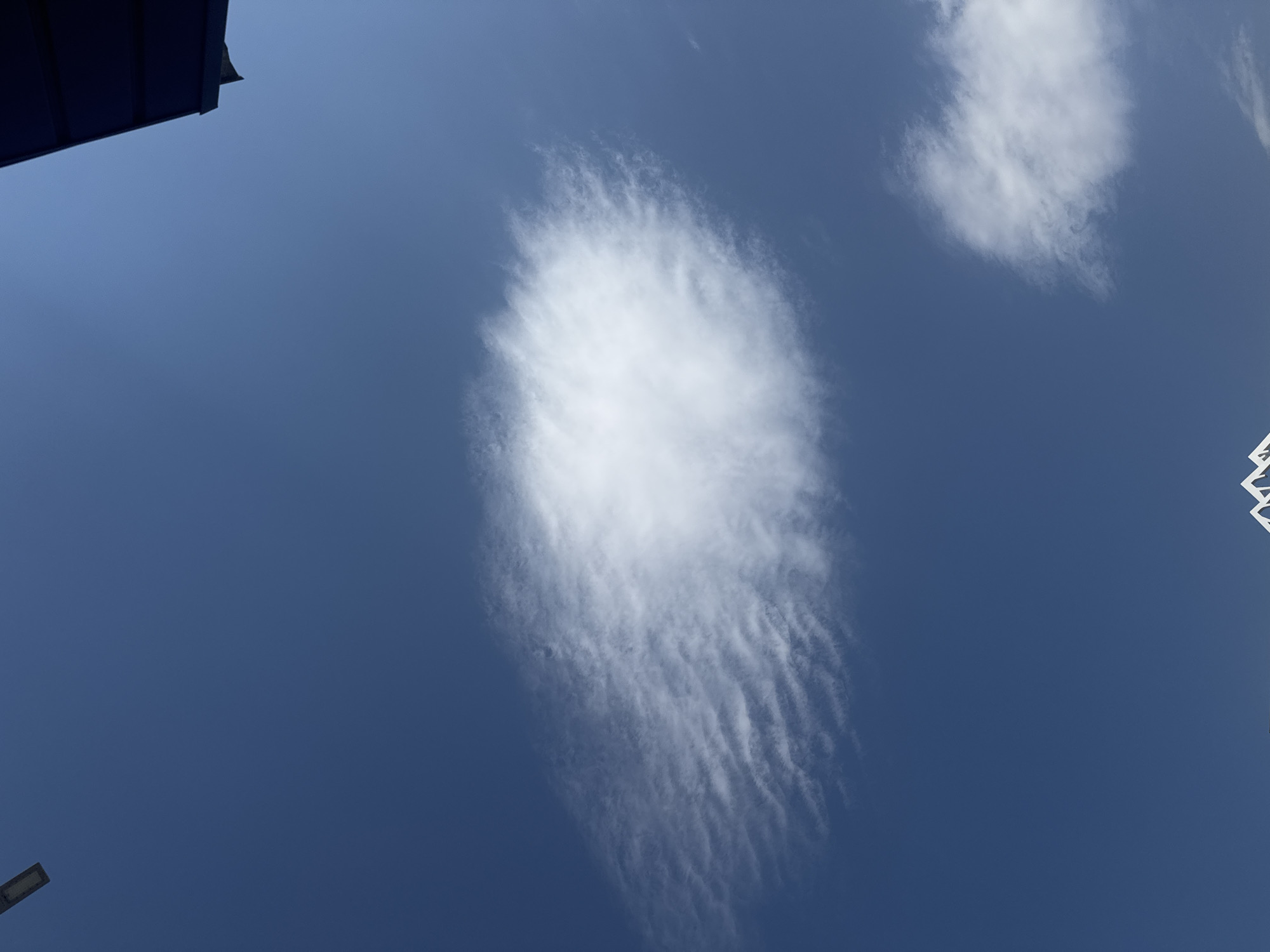 An outdoor, eye-level, wide shot captures a bright, clear blue sky with a few wispy clouds. In the upper left corner, a dark, rectangular object hangs from a pole, possibly a light fixture. Towards the upper right, a portion of a dark blue structure, possibly a roof or canopy, is visible. A large, elongated cloud with rippled texture stretches horizontally across the center of the frame, while a smaller, puffier cloud is situated in the lower right quadrant. At the bottom of the image, the white, angular peaks of a structure, likely a building, are just visible, hinting at an urban environment. The scene is consistent with a sunny day in Bishkek, Kyrgyzstan.
