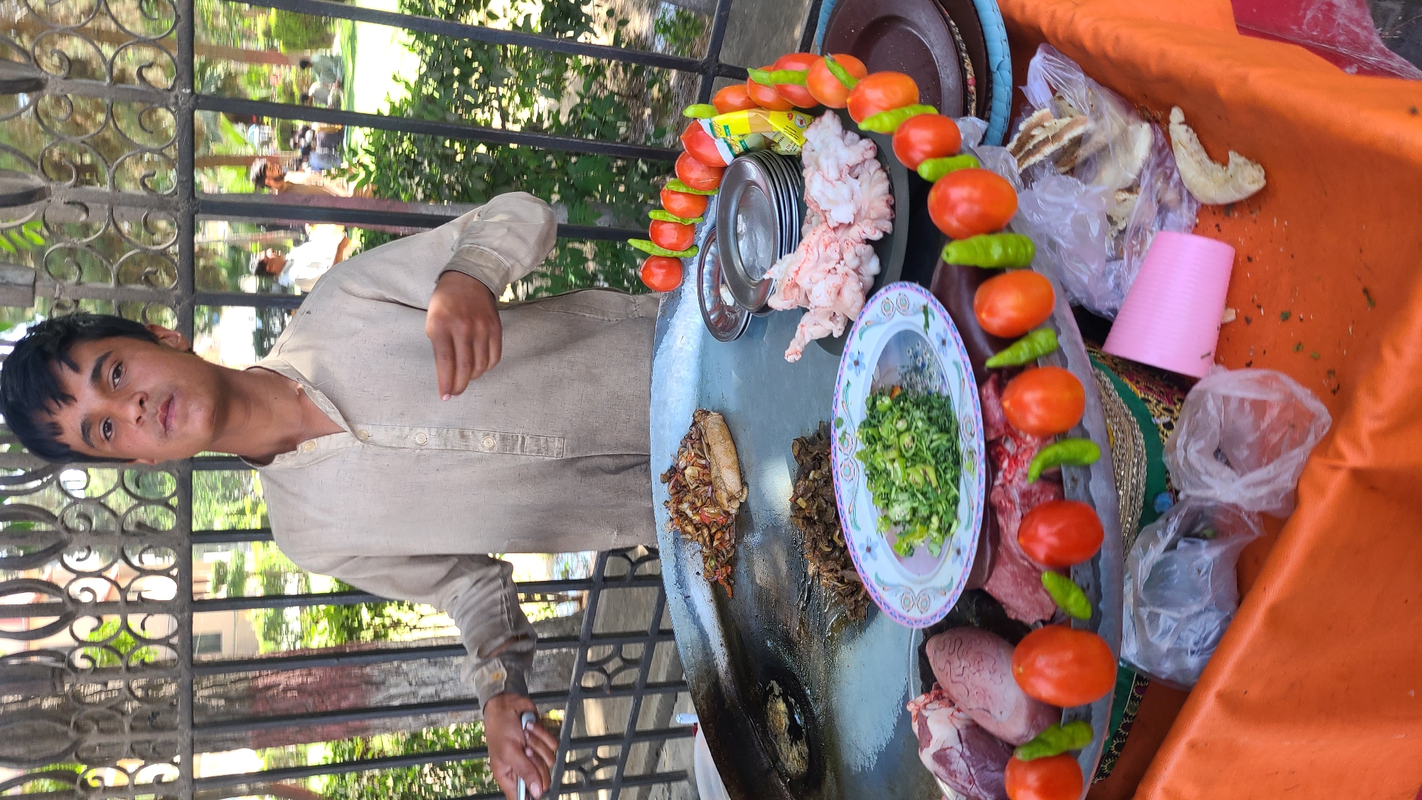A man stands next to a food cart in what appears to be a street food setting, likely in Quetta City Tehsil, Pakistan. He is positioned to the left of a large round cooking surface or griddle, where various food items are placed. These items include cooked meat pieces, raw meat, fresh vegetables in a bowl, and skewers of tomatoes and green chilies. Additional items, such as bread in plastic bags and a pink cup, are on a surface covered with orange fabric. The man is wearing a light-colored, button-down shirt. A black metal fence is visible behind him, with trees and foliage in the background.