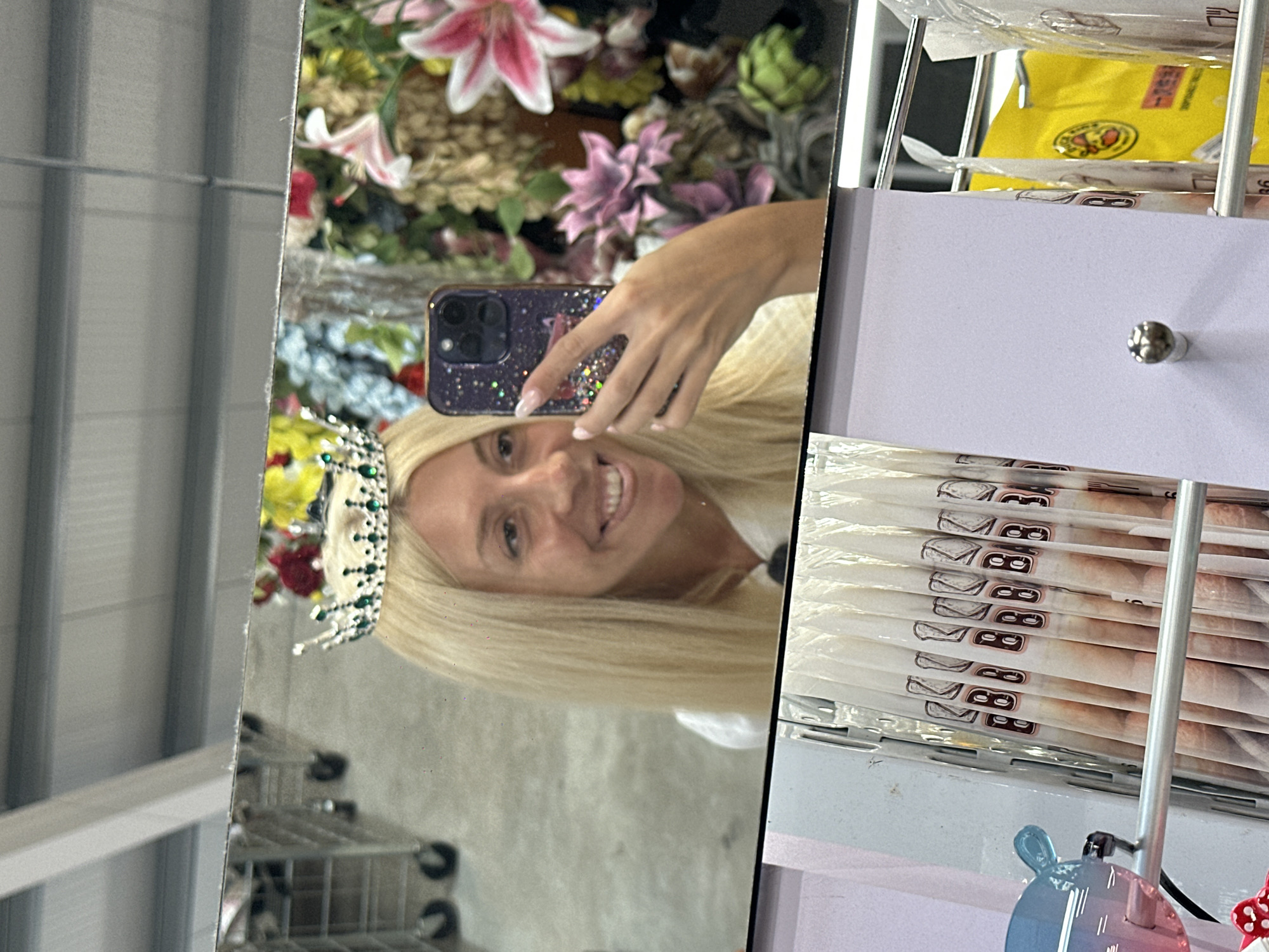 A woman with long blonde hair and a tiara is taking a selfie in a mirror. She is holding a phone in her right hand and smiling at the camera. The background is a display of artificial flowers. In the foreground, there is a rack of items with a numbered print on them. The scene appears to be indoors, possibly in a retail store. There is no information available about the location in Pazardzhik, Bulgaria.