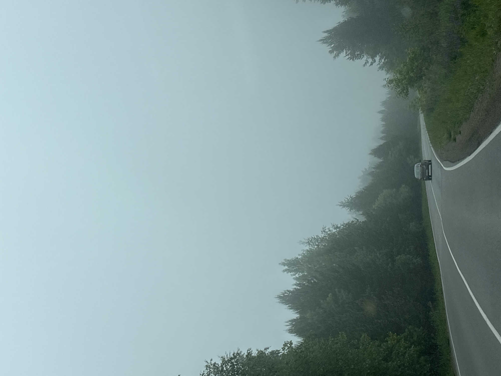 The image depicts a road scene in Chaikovsky, Russia, characterized by dense fog. A paved road stretches into the distance, its dark asphalt contrasting with the vibrant green of the trees and undergrowth lining both sides. A distinct solid white line marks the right edge of the road, while a dashed white line is partially visible towards the center, indicating lanes.
The most striking feature is the heavy, pervasive fog that blankets the entire environment, obscuring the horizon and significantly reducing visibility. The sky is completely enveloped by this light blue-gray mist, blending seamlessly with the distant landscape. The trees, though dark green and lush in the foreground, gradually fade into the thick fog, appearing as indistinct, ghostly silhouettes further down the road.
A dark-colored car, possibly an SUV, is visible ahead on the right side of the road, also navigating through the misty conditions. The photograph appears to be taken from inside a moving vehicle, looking forward, suggesting an activity of travel or driving. The diffused light suggests it is daytime, though the fog creates an overcast, soft illumination without clear shadows, contributing to a tranquil yet cautious atmosphere. There is no visible text in the image.