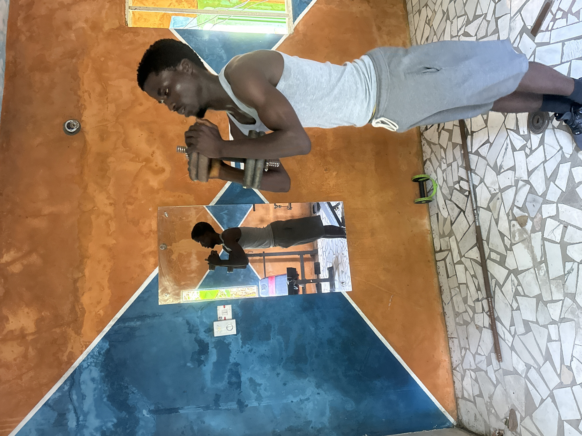 In Badagry, Nigeria, a young Black man is captured mid-workout in a room with a unique wall design. The walls are a striking combination of textured orange and a vibrant blue, separated by a diagonal white line. A mirror reflects the scene, showing the man from a different angle, along with a weight bench and a colorful stained-glass window. The floor is a mosaic of irregular white and grey tiles. The man, wearing a grey tank top and grey shorts, is holding a dumbbell with both hands, his arms bent as if performing a bicep curl. His reflection shows him in a similar pose, with a weight bench behind him. On the floor, a metal bar and a green ab roller are visible, suggesting a home gym setup. The room appears to be indoors, with natural light streaming in from the window.