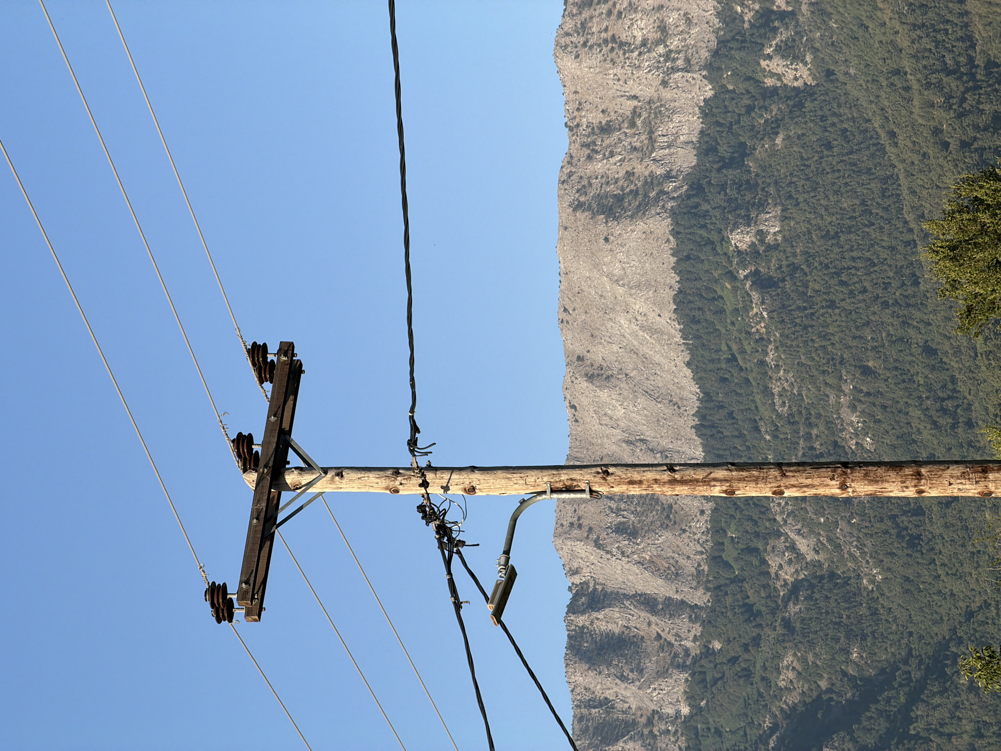 A wooden utility pole stands in the foreground, supporting multiple wires and a street lamp. The pole is weathered and appears to be made of rough-hewn timber. Attached to the top of the pole is a crossarm, also made of wood, from which several ceramic insulators are suspended. Wires are strung between these insulators, extending out of the frame. A street lamp is mounted on a metal arm extending from the pole, positioned below the crossarm. Behind the pole, a mountain range dominates the background, characterized by rocky slopes and dense green foliage, likely trees. The sky above is a clear, bright blue. The scene is likely located in a semi-rural or mountainous area.