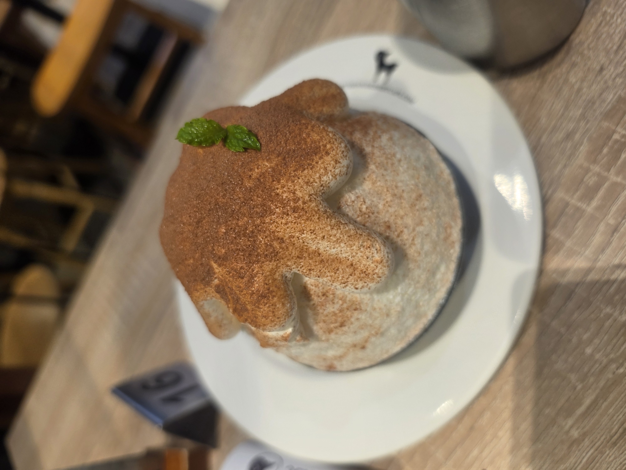 The image displays a close-up, high-angle shot of a dessert presented on a white plate with a dark animal silhouette logo, possibly a deer. The dessert itself is light-colored, likely cream or shaved ice, and features a distinct shape resembling an animal head, possibly a bear or a similar creature, covered in a generous dusting of brown cocoa powder. Two small, fresh green mint leaves garnish the top of the dessert, providing a vibrant color contrast.
The plate rests on a light-colored wooden table with a visible grain. In the blurred background, other elements of a cafe or restaurant setting are visible, including indistinct furniture and a metallic object, possibly a part of a drink container. A table number stand with the number "16" is partially visible in the lower left portion of the image, further confirming the restaurant context. The overall lighting suggests an indoor setting, possibly during the daytime. The focus is sharp on the dessert, with a shallow depth of field blurring the surroundings. The scene depicts a dessert ready to be consumed in a cafe in Bangkok, Thailand.