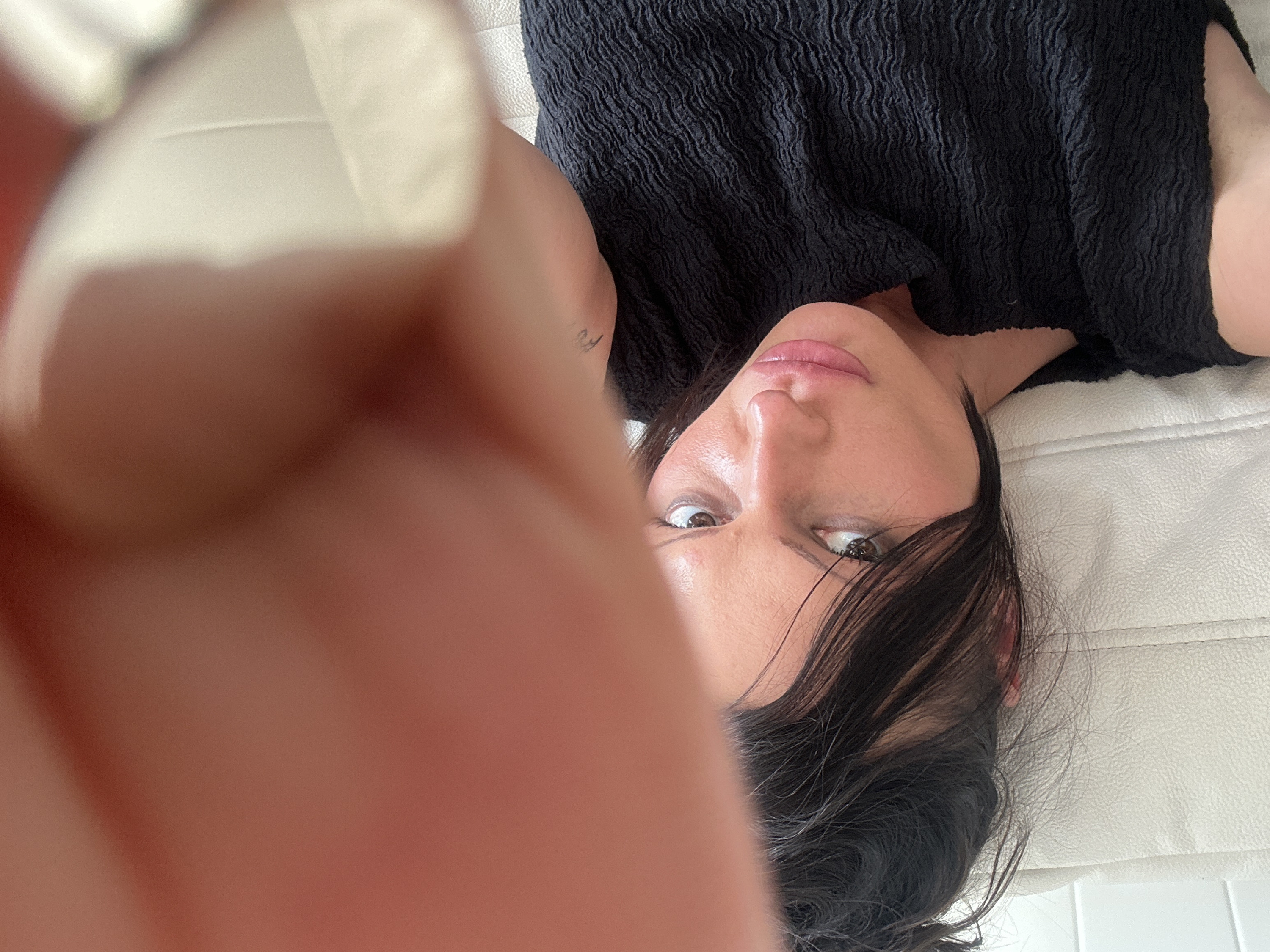 A close-up, high-angle interior shot features a female individual lying down and looking directly into the camera. A large, out-of-focus human finger occupies the upper portion of the frame, obscuring part of the scene. The woman has dark hair and brown eyes, with visible make-up, including pink lipstick. She is wearing a dark, textured garment with a rippled surface. A small, dark mark or tattoo is visible on her left arm. She is resting on a light-colored, possibly cream or white, upholstered surface with vertical stitching. To the far left, a white tiled-like surface is visible. There are no discernible visual cues within the image to identify a specific location in the city of London, United Kingdom.