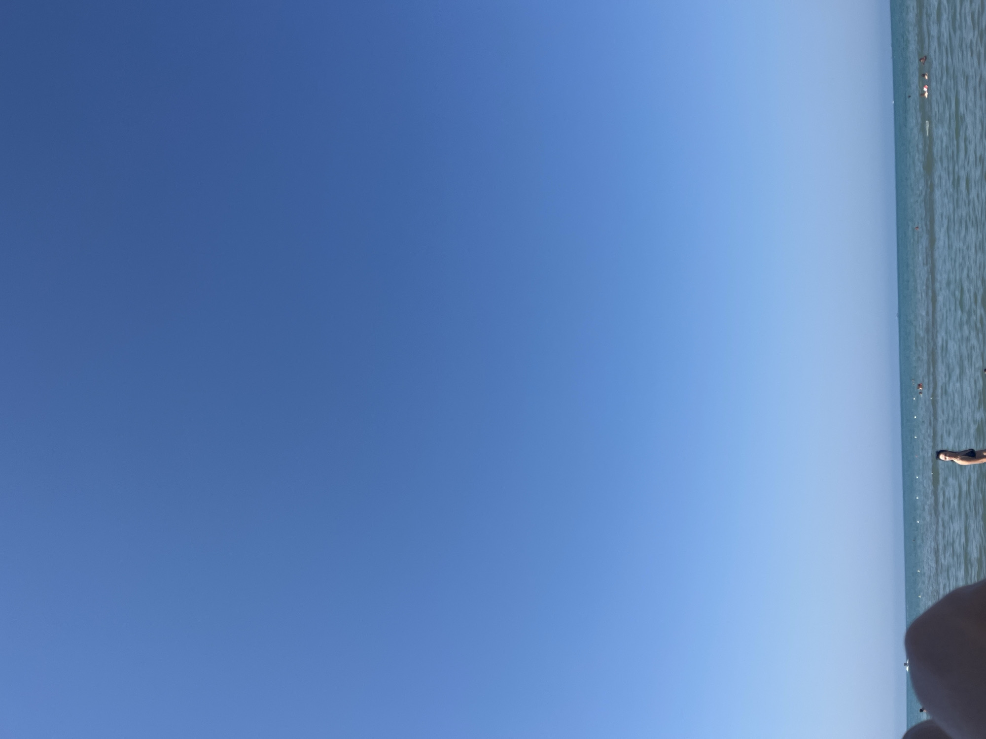 The image shows a clear blue sky over a calm sea, with a beach visible in the distance. Several people are in the water, swimming and enjoying the day. The perspective suggests the photo was taken from the beach, looking out towards the sea. The location is identified as the city of Bashkia Durrës, Albania. The overall scene is one of a typical summer day at the beach.