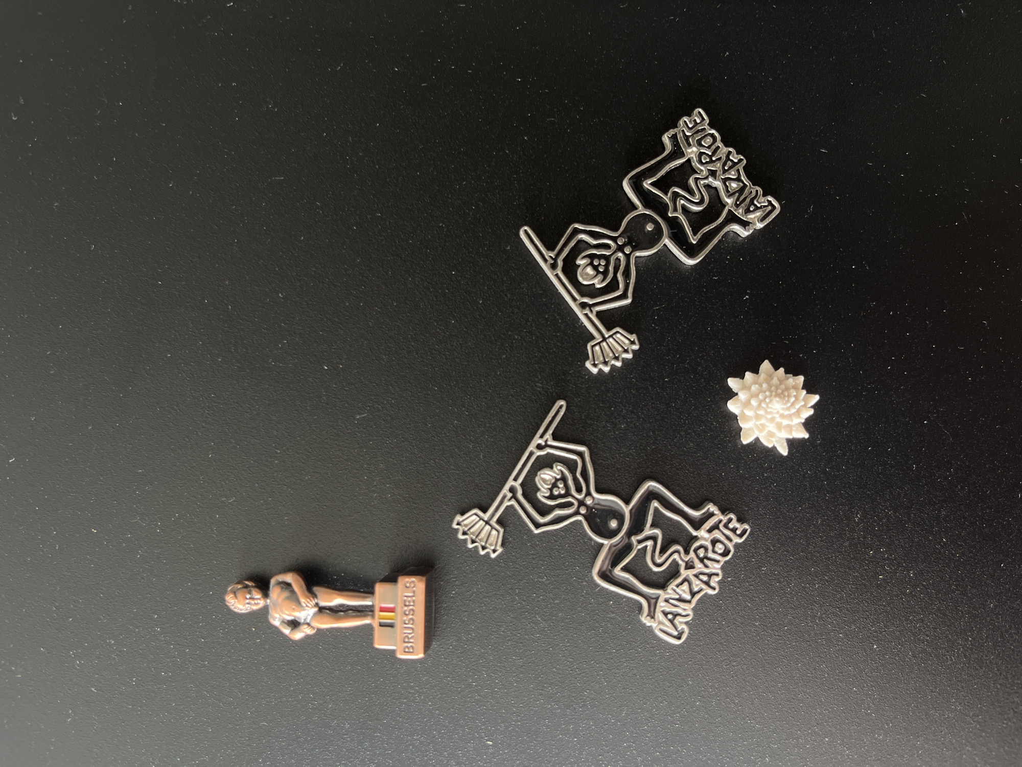 A small copper figurine of the Manneken Pis, a landmark in Brussels, Belgium, stands on a pedestal with the word "BRUSSELS" and the Belgian flag printed on it. To the right of the figurine are two silver-colored metal charms depicting a stylized human figure holding a broom, with the word "LANZAROTE" integrated into its design. In the lower center of the image is a small, intricate white decorative object resembling a flower. The objects are arranged on a plain, dark, textured surface, possibly a table. The lighting suggests an indoor environment. There is no indication of the city of Moscow, Russia, in the image.