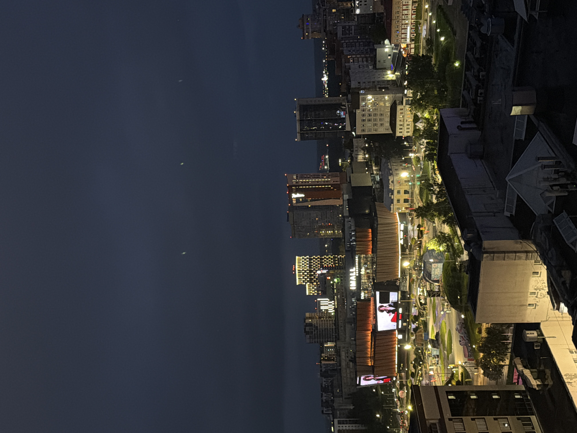 This is a nighttime aerial shot of a city scene, likely in Perm, Russia, focusing on illuminated buildings and urban infrastructure. The foreground features the dark, flat rooftops of several buildings, some with visible ventilation units. Beyond these, a mid-ground shows a public space with green areas, trees, and pathways, illuminated by numerous streetlights. Prominently featured are large buildings with illuminated facades and extensive digital billboards displaying advertisements. One billboard shows a person, likely a model or presenter. Further in the background, a cluster of taller buildings, including skyscrapers, are visible. Some of these taller buildings have intricate patterns of light on their facades, suggesting modern architectural design. The sky is dark and overcast, typical of evening or night in a city. The overall impression is of a bustling, illuminated urban environment at night.