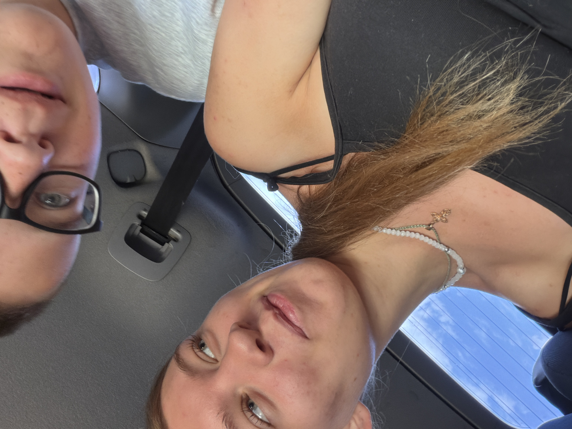 Two individuals are in a vehicle, their faces captured in a close-up, overhead shot. One person, in the foreground, is a young female with light brown hair, wearing a black tank top and a multi-layered beaded necklace with a small, gold-colored charm. Her gaze is directed upwards and to the left, with a slight smile. The other individual, partially visible in the upper portion of the image, appears to be wearing glasses and a light gray shirt. The vehicle's interior is visible, including a seatbelt and the textured surface of the car's headliner. The background outside the car shows a bright blue sky with scattered clouds, suggesting it is daytime. There is no indication of the specific location within Rostov-on-Don, Russia.
