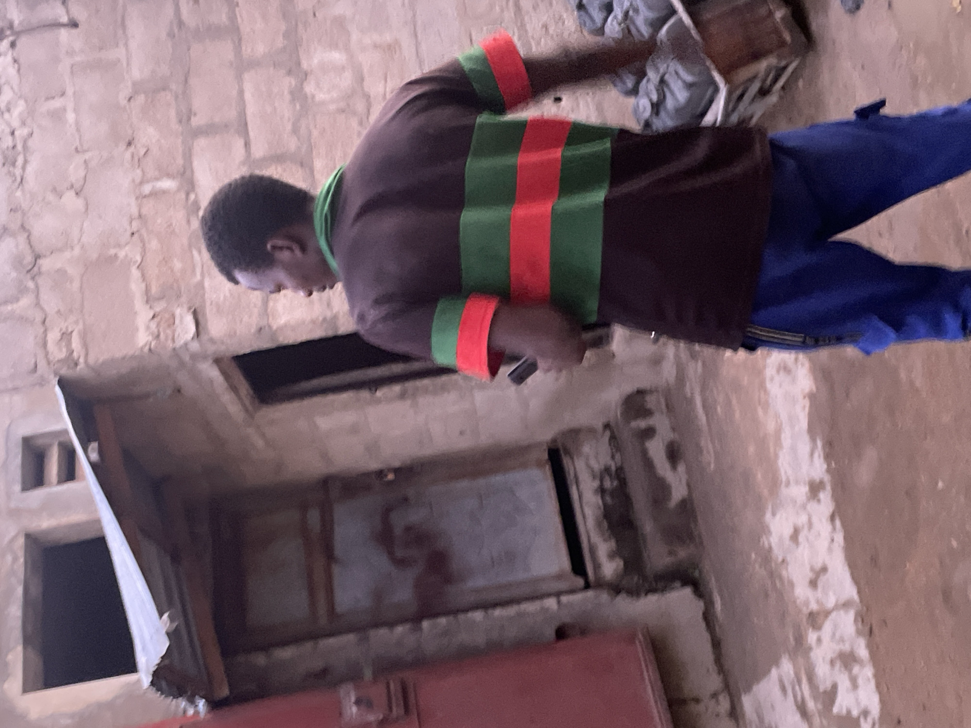 The image, rotated clockwise, captures a person standing in what appears to be an indoor or semi-enclosed space with rough, light-colored brick walls. The ground is a mix of dirt and concrete.
The subject is a person, likely male, with short dark hair, wearing a dark brown long-sleeved shirt with a green collar. The shirt features prominent horizontal green and red stripes across the torso and on the cuffs. He is also wearing blue trousers. The person is seen looking downwards, and is holding a dark-colored cell phone in their right hand.
In the background, to the right of the person, there's a wooden crate or box, partially obscured by some grey fabric or material. Towards the lower left, there is a large, dark rectangular opening in the wall, possibly a doorway or an access hatch, leading into a darker interior. Above this opening, another smaller square opening or vent is visible. A red rectangular object, possibly part of a door or container, is present in the lower foreground.
The lighting suggests daytime, but it's not possible to determine the exact time or weather conditions. No specific emotions are discernible on the person's face, which is mostly turned away. There is no visible text in the image.