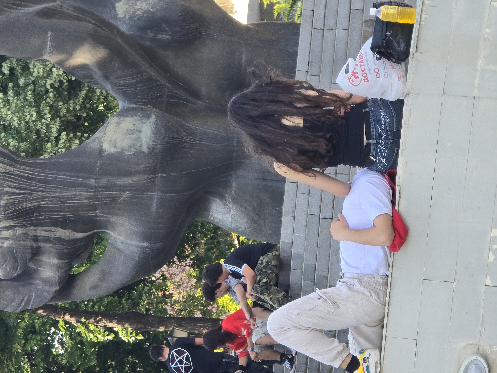 Observed in an outdoor setting, possibly a park, is a stone sculpture next to a group of people. A woman with long, dark hair stands near the sculpture, while a man in a white t-shirt and light-colored pants is seated nearby. Other individuals, including one wearing a black shirt with a pentagram, are situated in the background. Additional details include a plastic bag labeled "ДОСТАВ" and bottles nearby. The location appears to be in the city of Unknown, Unknown.