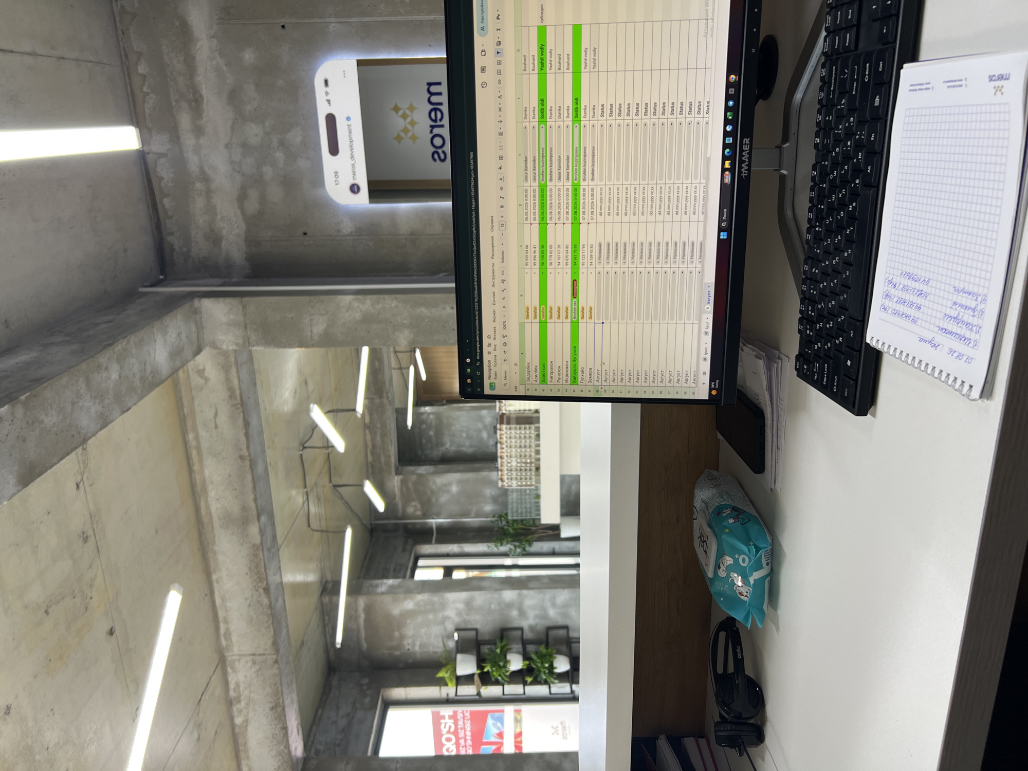 The image captures an office interior that appears to be in the city of Shahrixon, Uzbekistan, given the Cyrillic script visible on the screen and in the notebook. The scene is dominated by a modern, somewhat minimalist aesthetic with exposed concrete walls and ceilings, along with fluorescent tube lighting.
In the foreground, a white desk holds a computer monitor displaying a spreadsheet, a black keyboard, and a spiral-bound notebook with handwritten notes. To the left of the monitor, a pair of black headphones and a package of wet wipes are visible. A smartphone rests on a stack of papers to the right of the wet wipes. The spreadsheet on the screen contains columns with what seem to be names, dates, and status updates, suggesting an administrative or booking system. The notebook contains a list of items with associated numbers and dates, possibly related to tasks or appointments.
The background reveals a reception area or office lobby with large windows, plants in pots on shelves, and what appears to be a miniature architectural model or display. A framed digital advertisement or information screen displaying the "meros_development" logo and the time "17:09" is mounted on the concrete wall. The overall impression is of a contemporary business environment.