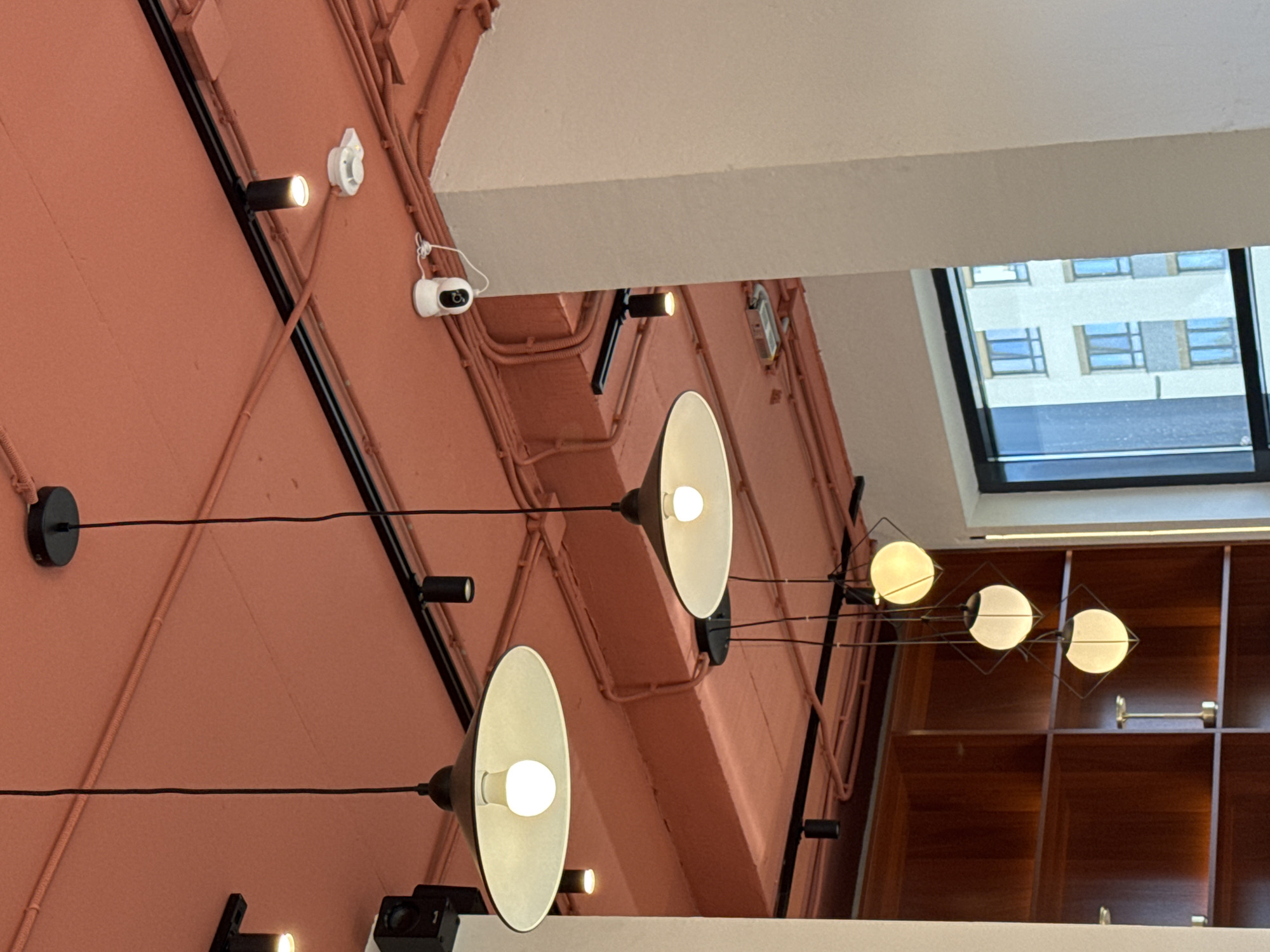 The image shows a collection of ceiling lights and security cameras mounted on a pink wall. The lights vary in design, with some being track lighting with small, cylindrical fixtures, while others are pendant lamps with wide, conical shades. One set of pendant lamps features multiple spherical bulbs attached to a wire frame. A white security camera with a lens and a small white base is visible on the upper left side of the wall. Another small, white circular device, possibly a smoke detector or another sensor, is mounted above it. The wall is covered in electrical conduits and wiring, suggesting an interior space with exposed infrastructure.
In the background, a window reveals a glimpse of an urban environment with a building featuring multiple windows. The time of day appears to be daytime, as indicated by the natural light entering through the window. The overall impression is of a modern interior space, possibly an office, retail store, or public building, that incorporates industrial design elements. There is no visible text in the image.