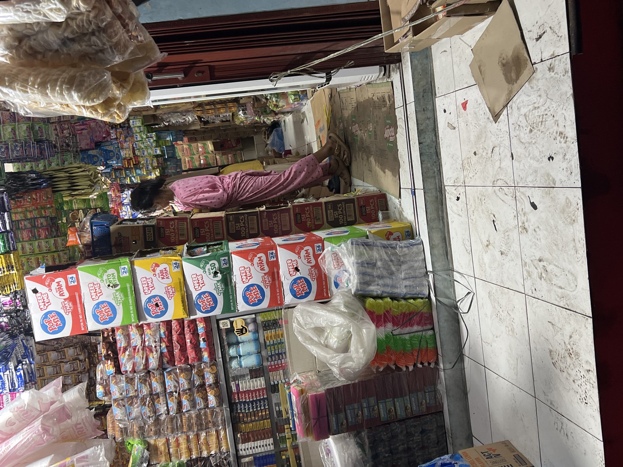 This is a medium shot of a small grocery store in Citeureup, Indonesia. A young woman in a pink pajama set is standing in the aisle, looking at a display of instant noodle boxes. The store is packed with merchandise, with shelves filled with packaged goods, snacks, and drinks. The floor is tiled and appears to be dirty with footprints. The overall impression is that of a typical, cluttered Indonesian convenience store.