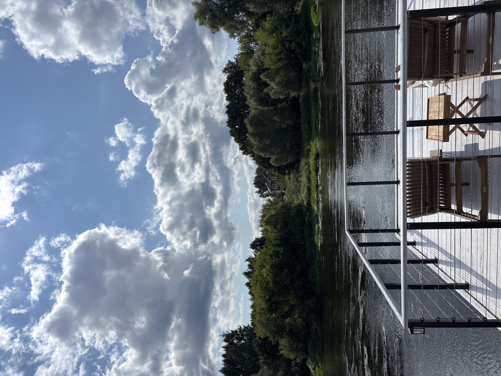 The image depicts a serene view from a wooden deck overlooking a wide, dark river. The deck is furnished with two empty wooden lounge chairs and a small wooden side table. In the distance, across the river, a lush, green treeline dominates the landscape, with a small house visible peeking through the foliage. The sky above is a vibrant blue, adorned with dramatic, fluffy white clouds, suggesting a pleasant, possibly warm day. The overall atmosphere is tranquil and inviting. This scene is likely located on the Moskva River, near the town of Zvenigorod, Russia, known for its scenic beauty and proximity to natural landscapes.