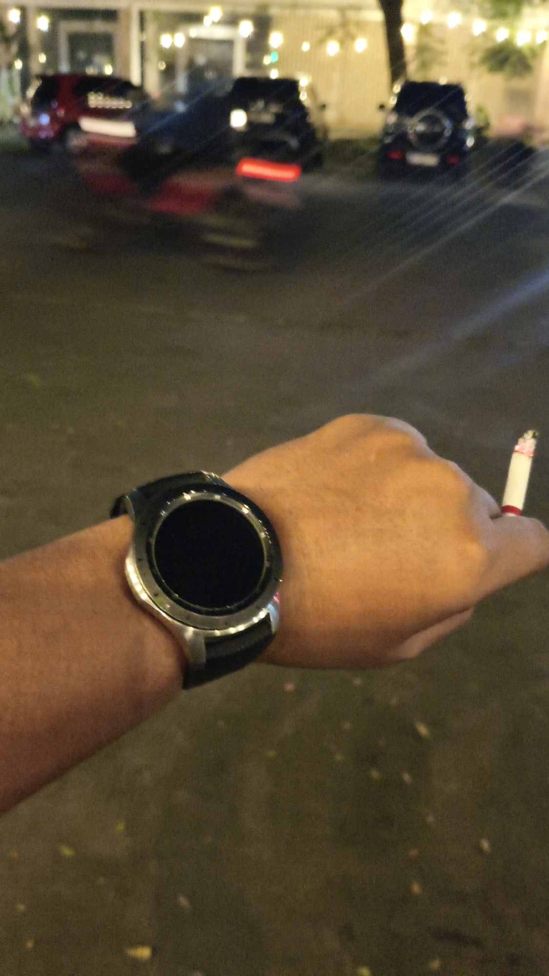 The image captures a close-up view of a person's left hand wearing a black and silver smartwatch. The person is holding a lit cigarette between their index and middle fingers. The background is blurred, showing a street scene at night with several cars parked. In the distance, there are buildings with illuminated windows and string lights, suggesting an urban environment. The presence of cars and buildings indicates a street in Karangsatria, Indonesia. The overall lighting suggests it is nighttime or dusk, with artificial lights from the buildings and cars illuminating the scene.
