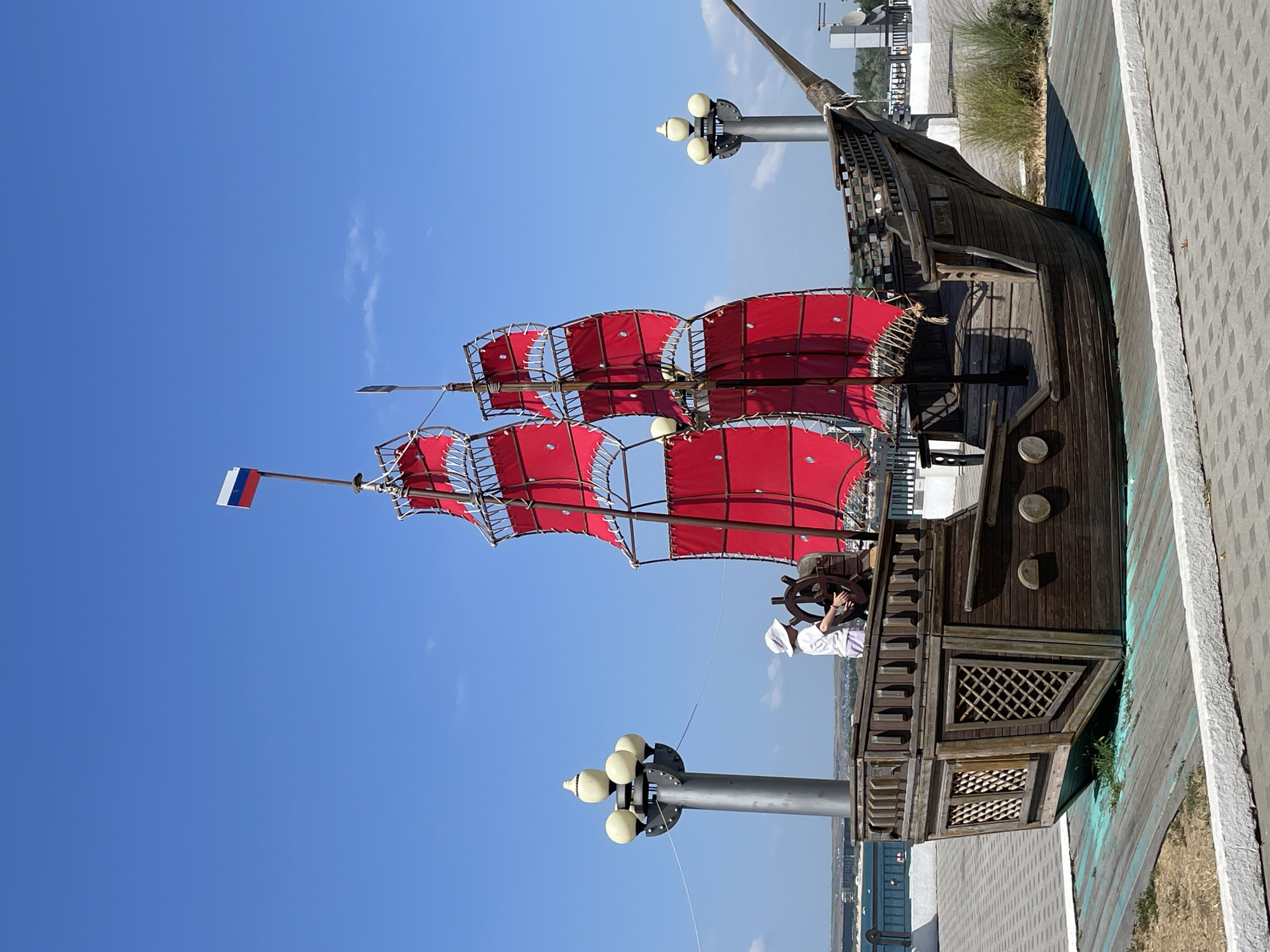 A wooden pirate ship replica with bright red sails is displayed outdoors on a sunny day, with a clear blue sky overhead. A person wearing a white hat and shirt stands at the ship's helm, appearing to steer. The ship is decorated with a Russian flag at its mast. The vessel is situated on a paved area with decorative lampposts on either side, and a body of water can be seen in the background. This decorative ship is located in the city of Anapa, Russia, likely on its waterfront promenade or a public park area.