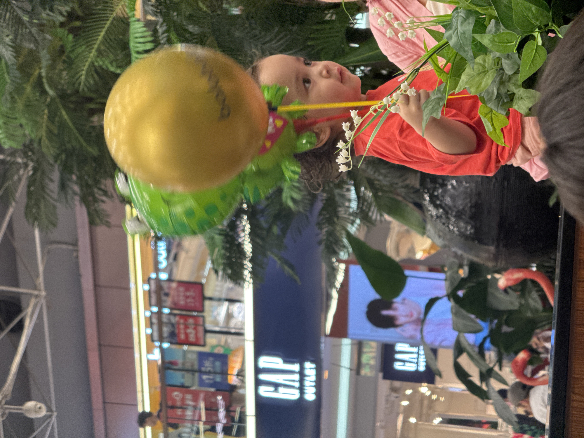 The image depicts a young child in a red shirt, holding a lily of the valley flower. The child also holds a yellow balloon. The background reveals a shopping mall environment. "GAP OUTLET" is visibly written on a sign. The location appears to be in Haizhu District, China.