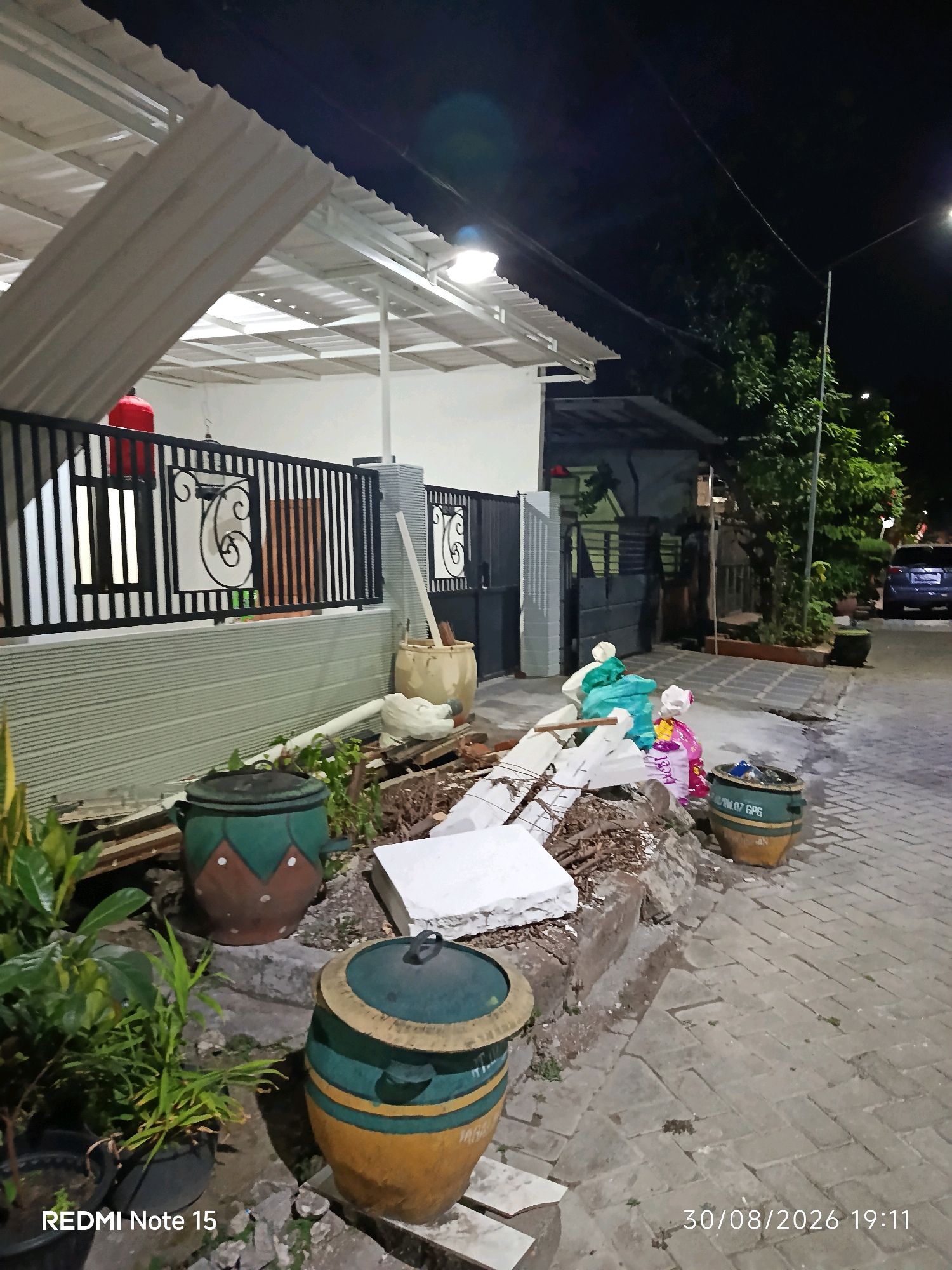 At night in Keboananom, Indonesia, a residential property is shown, with construction materials and refuse scattered near the front gate and sidewalk. A light fixture is visible on the roof overhang, illuminating the area. The property features a black metal fence with decorative scrollwork and a portion of a white corrugated metal roof. Two large, decorative ceramic pots are present; one is green and brown, and the other is yellow and green. Several plastic bags filled with debris are piled near the pots. A blue car is parked on the paved street in the background. The image is dated August 30, 2026, at 7:11 PM.
