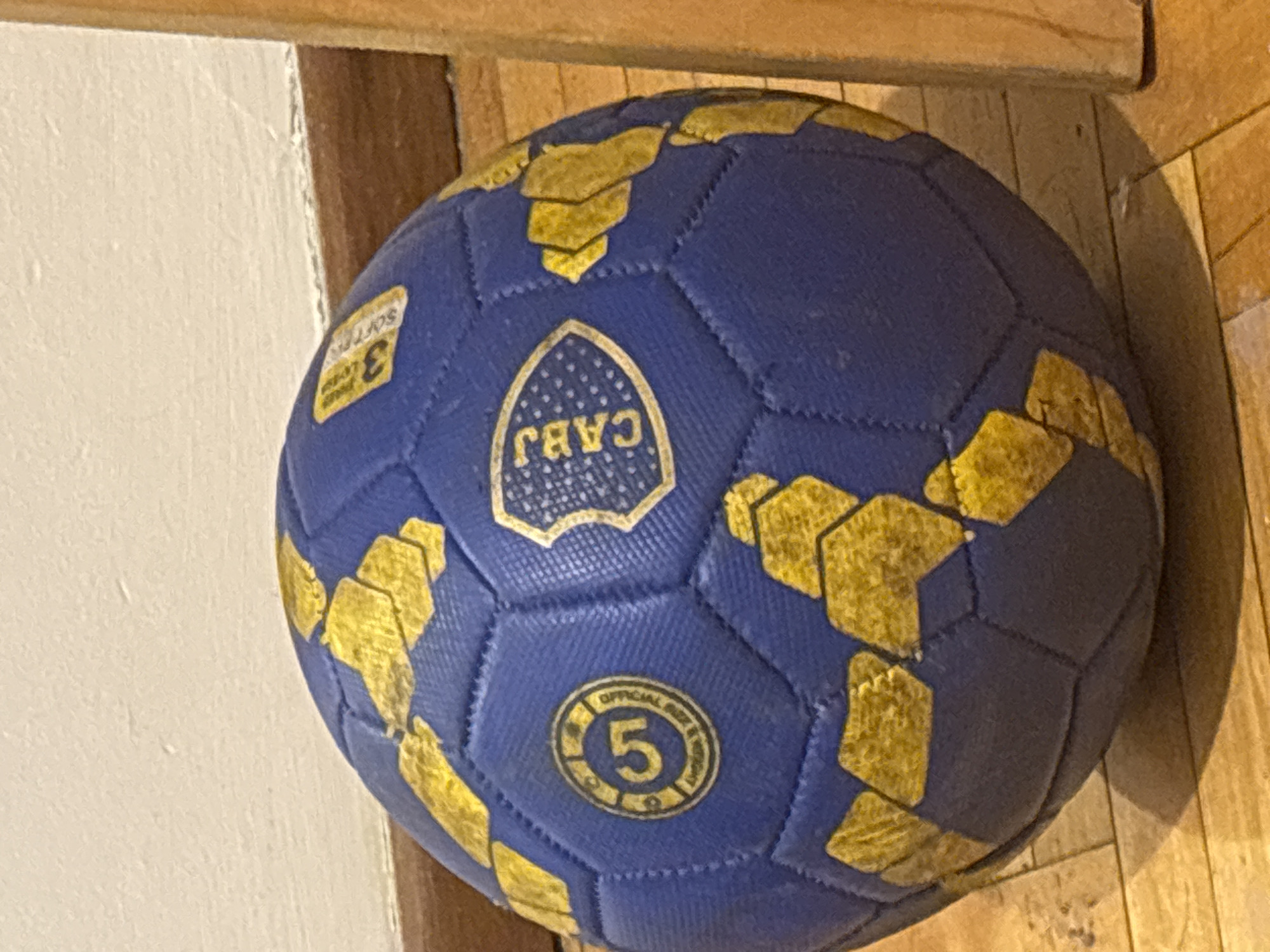 A blue soccer ball with gold-colored markings rests against a white wall and wooden floor. The soccer ball features a logo with the text "CABJ" and a number "5" inside a circular emblem, indicating its association with the Club Atlético Boca Juniors, a soccer team from Buenos Aires, Argentina. It also includes a "3 Layers Soft" sticker on one of its panels.