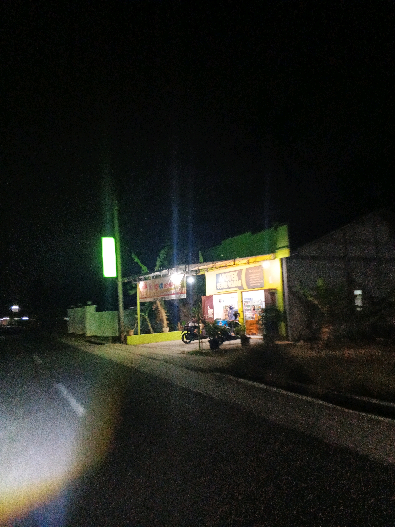 The image depicts a nighttime scene on a street in Sirau, Indonesia, featuring a brightly lit pharmacy. A green illuminated sign stands to the left of a building facade that is painted green and yellow. The pharmacy, identified by the sign "APOTEK SEVEN WARAS," has its interior illuminated, revealing shelves stocked with products and at least two individuals inside. In front of the pharmacy, two motorcycles are parked. The road in the foreground is dark, with a prominent light glare from what appears to be a vehicle's headlights, casting a rainbow effect on the asphalt. The overall environment suggests a commercial area open late at night.