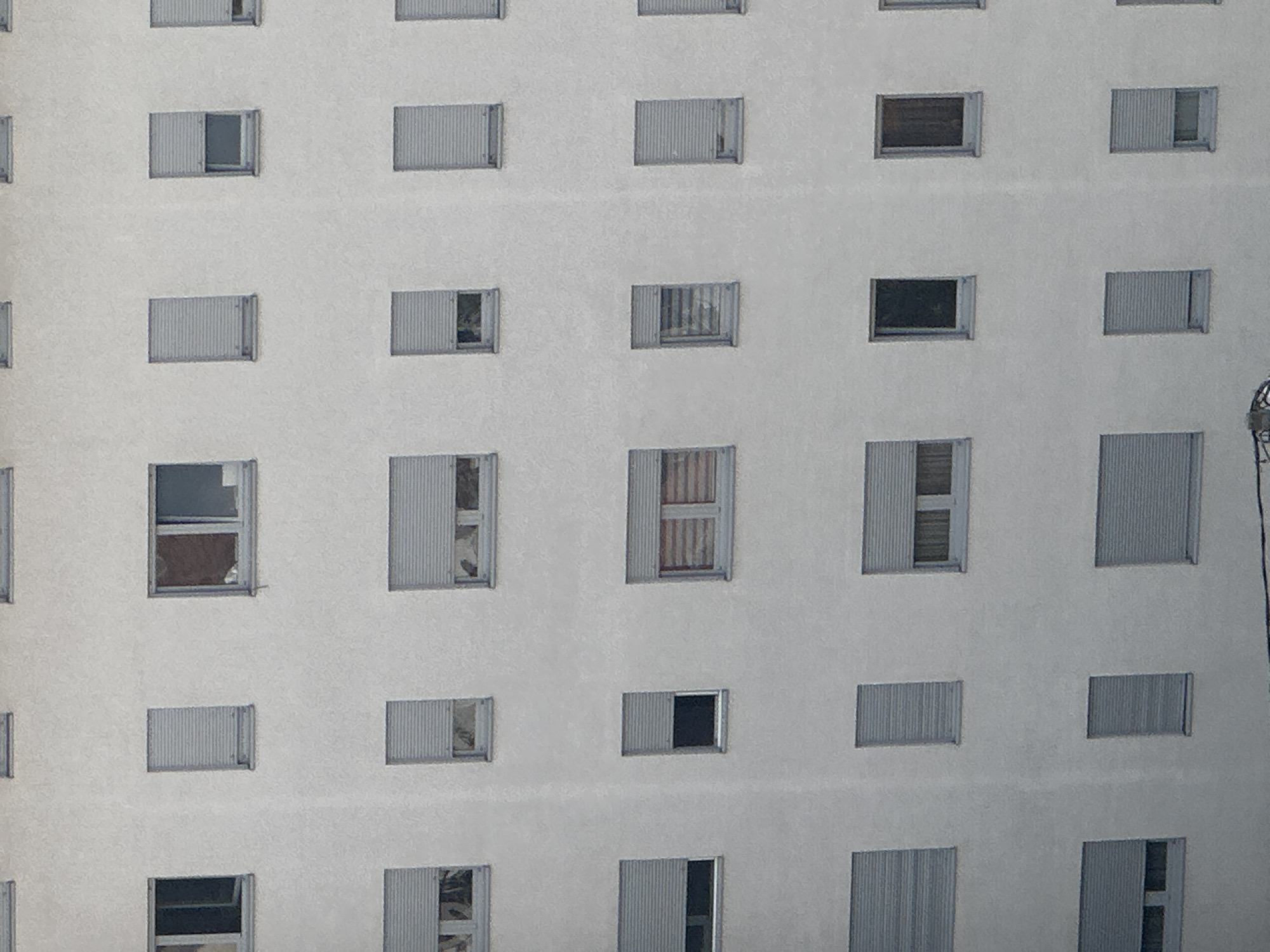 This is a close-up, eye-level, exterior, medium shot of a modern apartment building's white, textured facade. The building features a grid-like pattern of numerous uniformly sized windows, many of which are covered by grey roller shutters, some partially lowered, others fully closed, obscuring the interior views. A few windows show glimpses of interior blinds or subtle hints of what might be within. The overall impression is one of uniformity and modern architecture. The location in Belgrade, Serbia cannot be determined from the image.
